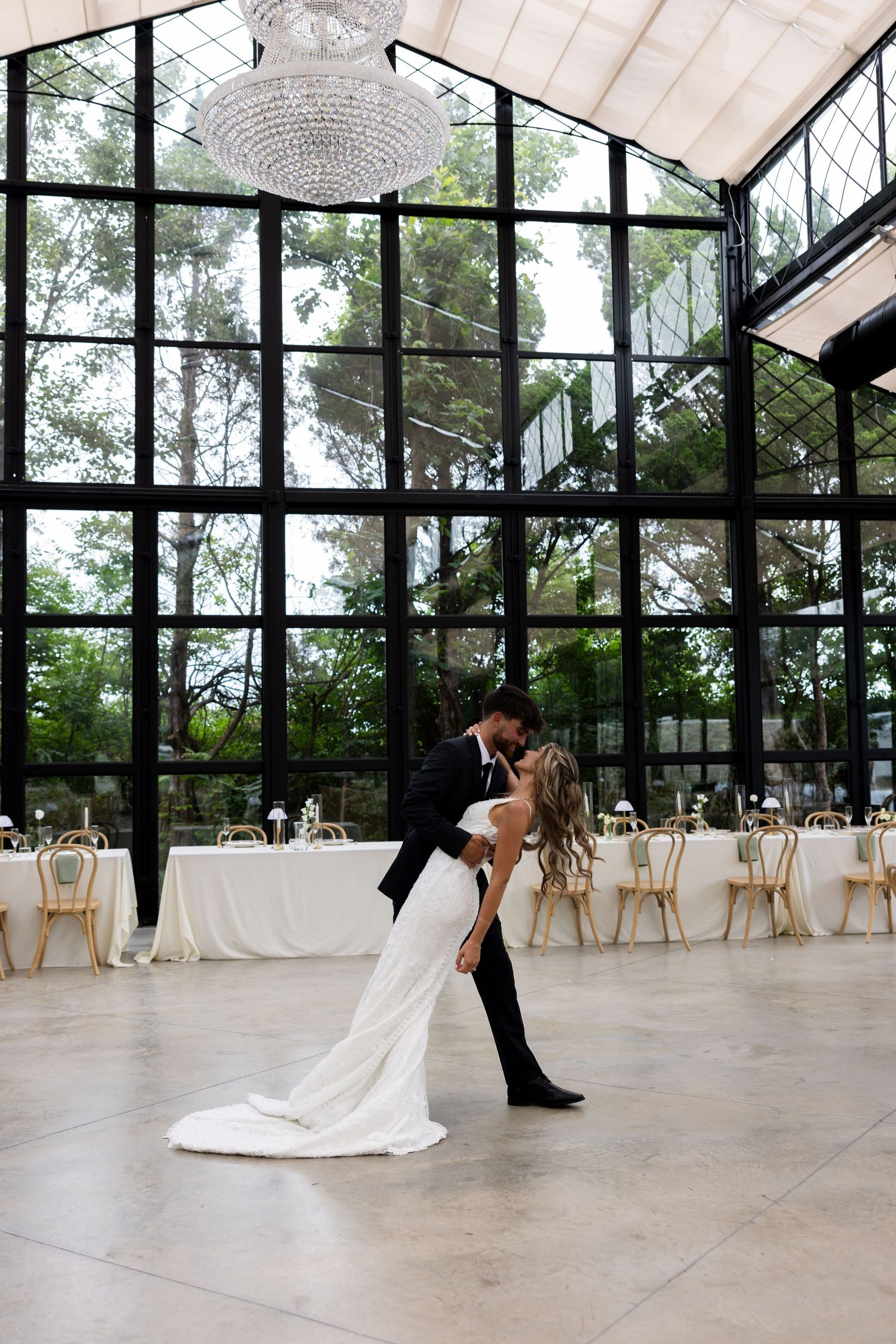 Bride and groom share romantic first dance dip in elegant greenhouse wedding venue with chandelier and floor-to-ceiling windows.