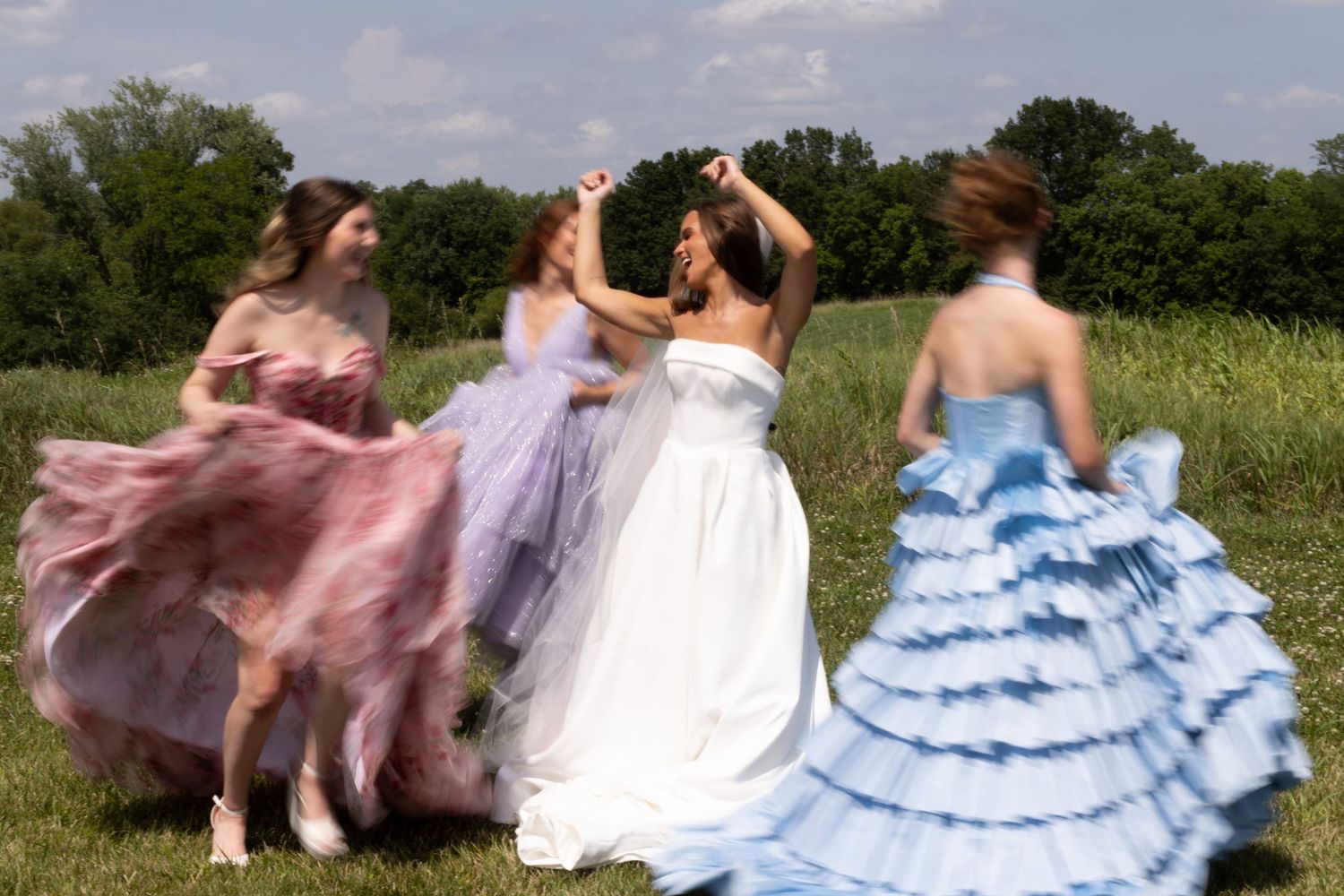 Four people in flowing formal dresses twirl and dance together in a sunny meadow with trees in the background.