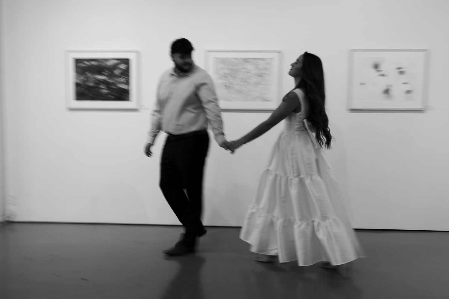 A couple in formal attire dances together in an art gallery with framed artwork displayed on white walls.