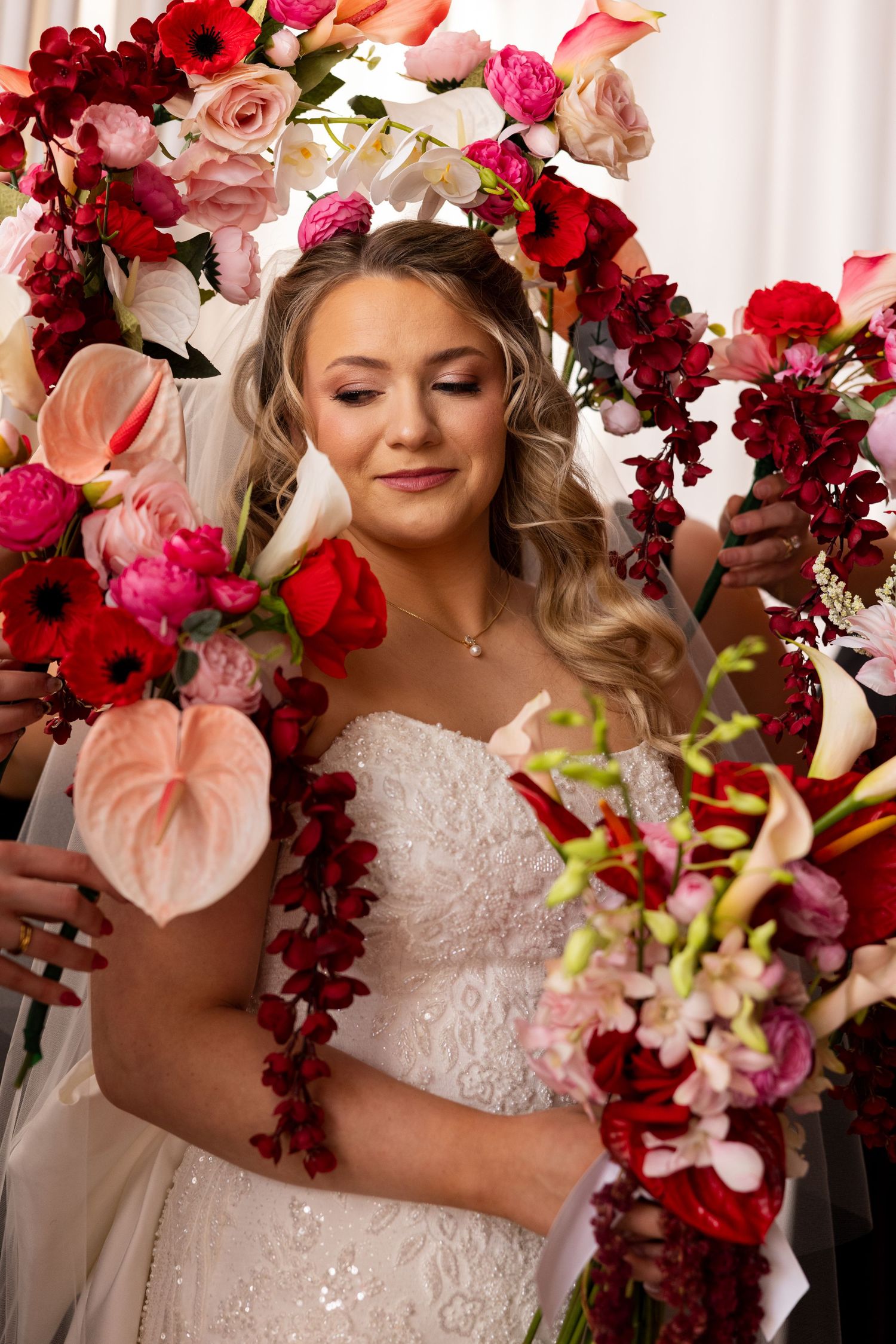 Bride in white lace gown holding red and pink floral bouquet, surrounded by vibrant flower arch on wedding day.