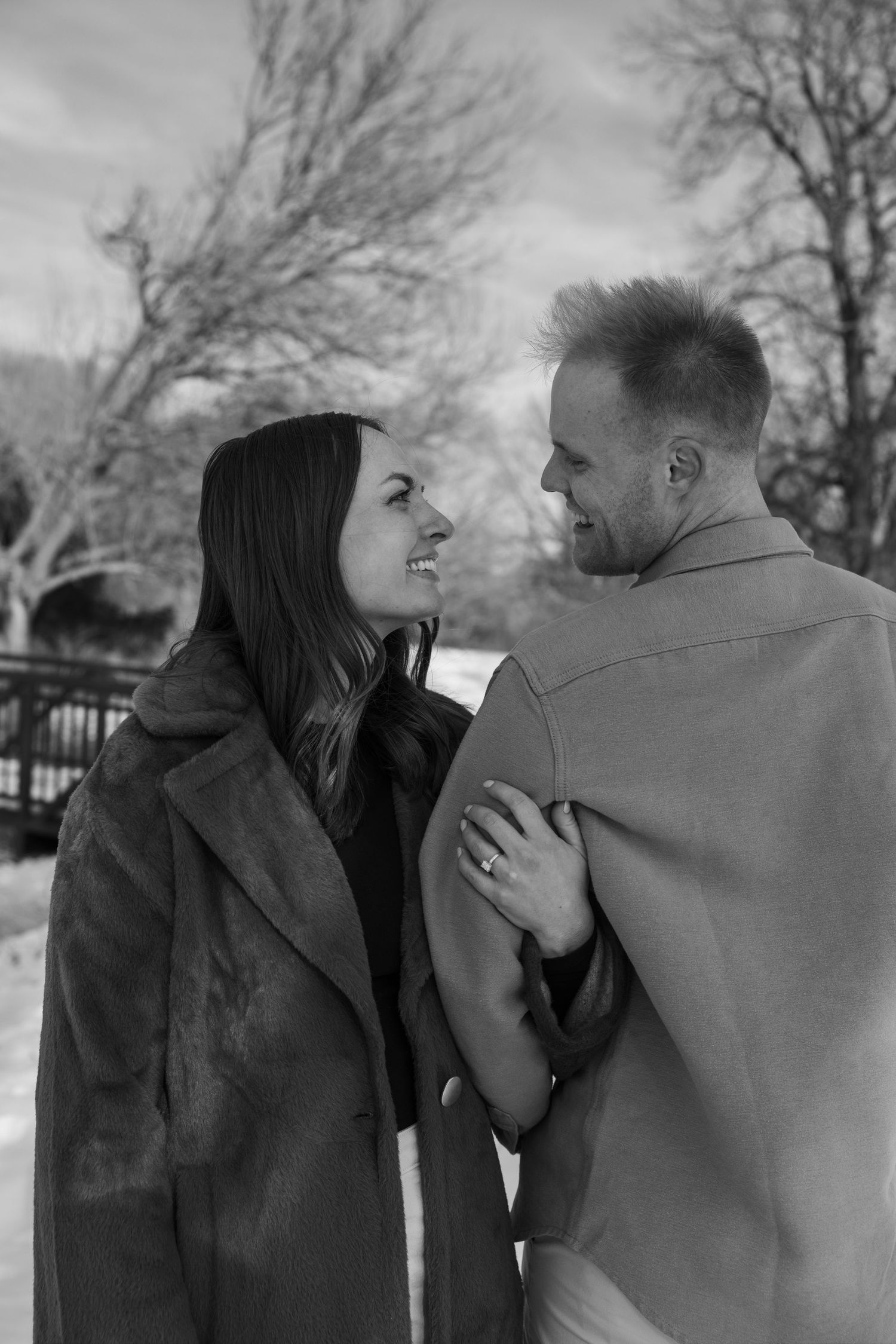 Black and white photo of a smiling couple gazing at each other outdoors in winter, woman in fur coat holding mans shoulder.