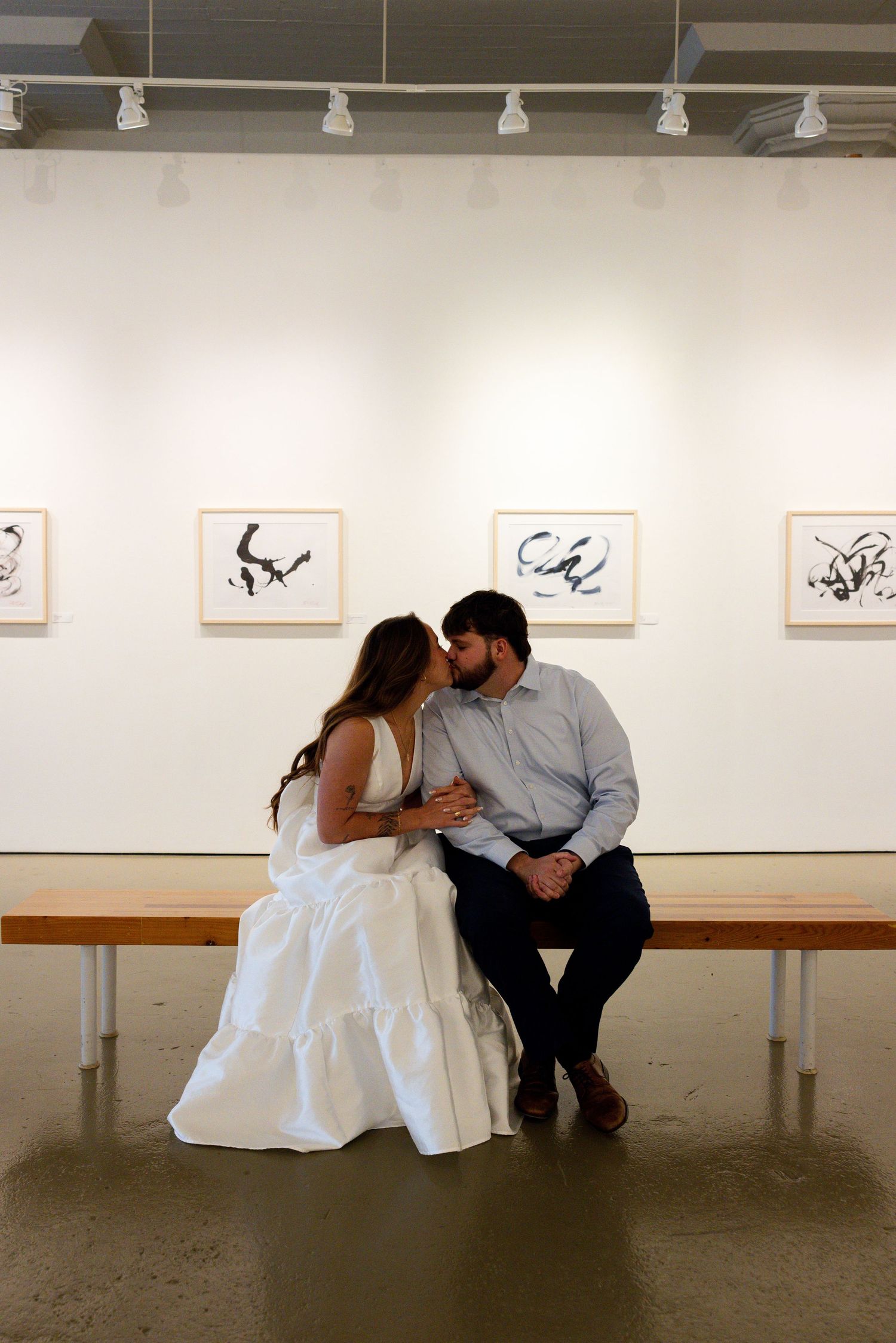 A couple shares an intimate moment while sitting on a bench in an art gallery with framed artwork on display.