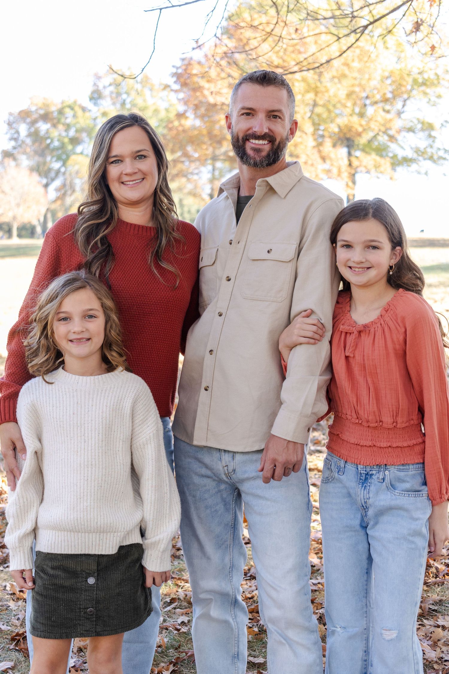 A family of four poses together outdoors during autumn wearing coordinated casual outfits.