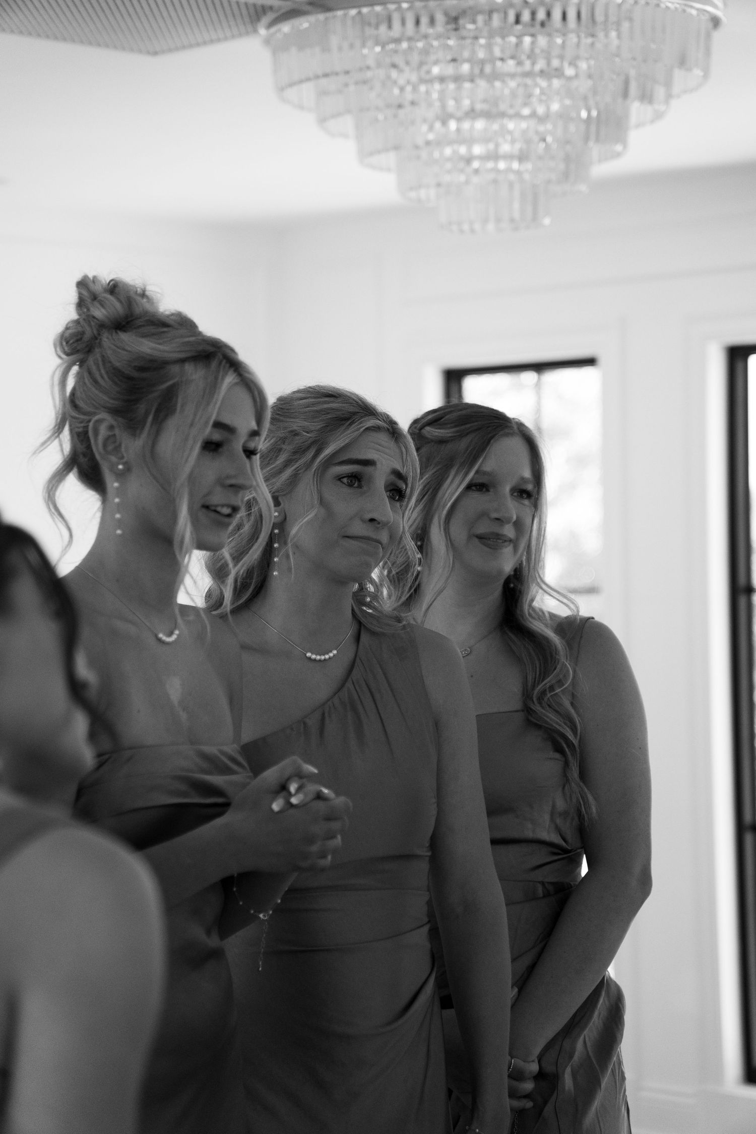 Black and white photo of emotional bridesmaids in matching dresses reacting to seeing the bride, elegant chandelier above.