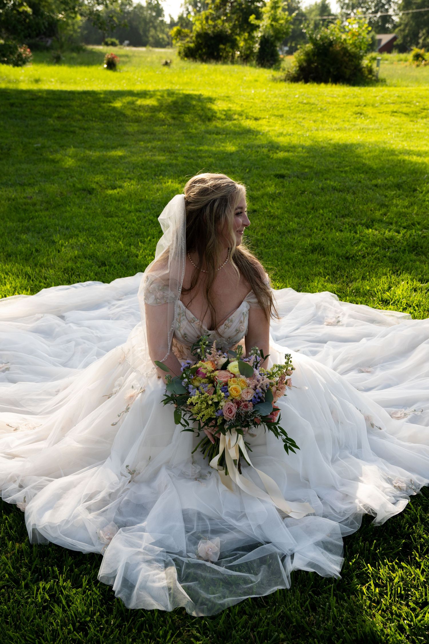 Bride in a flowing white tulle gown sits on green grass, smiling while holding a colorful bouquet with veil cascading behind.