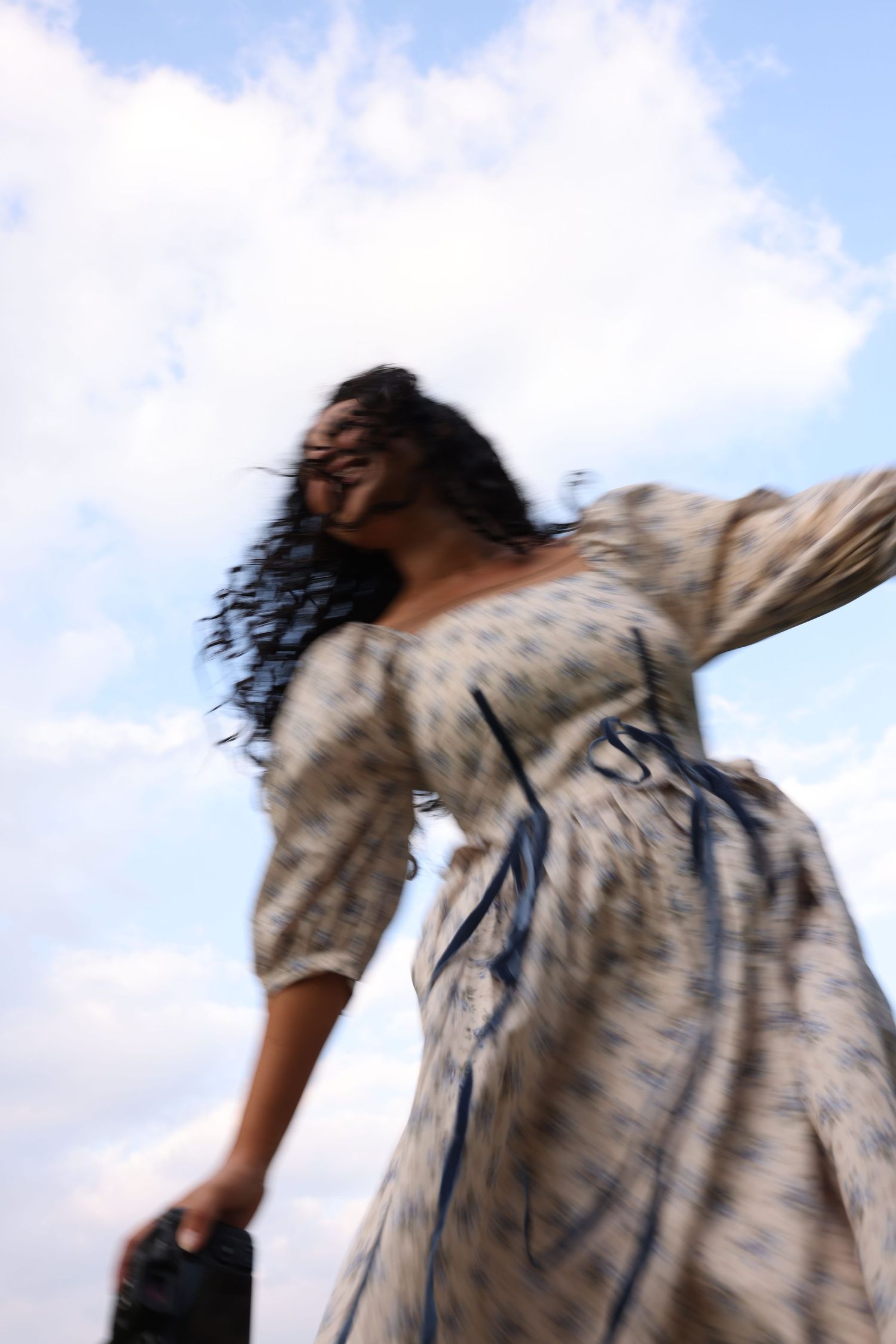 Woman in floral dress spinning outdoors against a cloudy blue sky, hair flowing in motion blur.