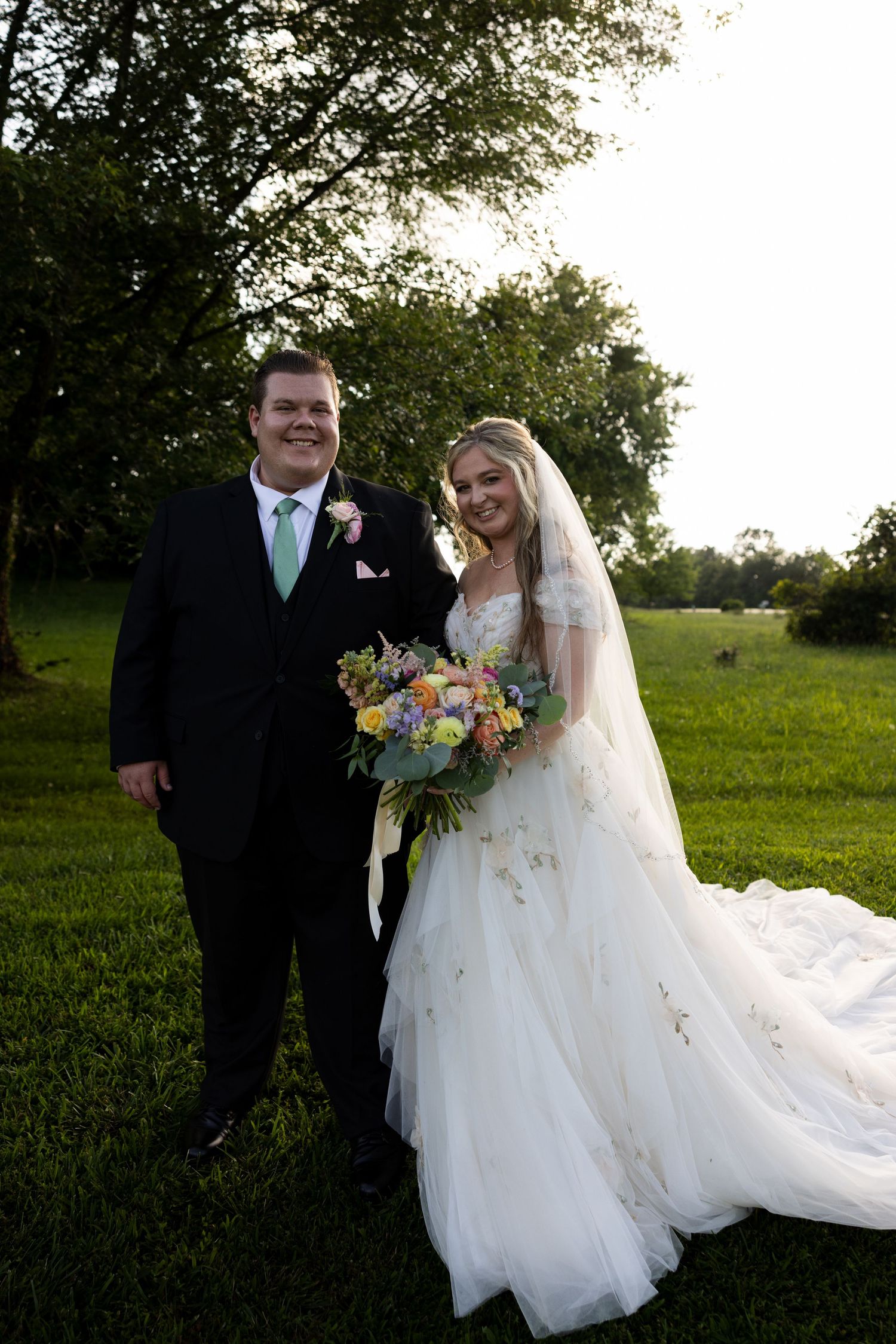 Bride in white gown holding colorful bouquet stands with groom in black suit on green lawn at golden hour.