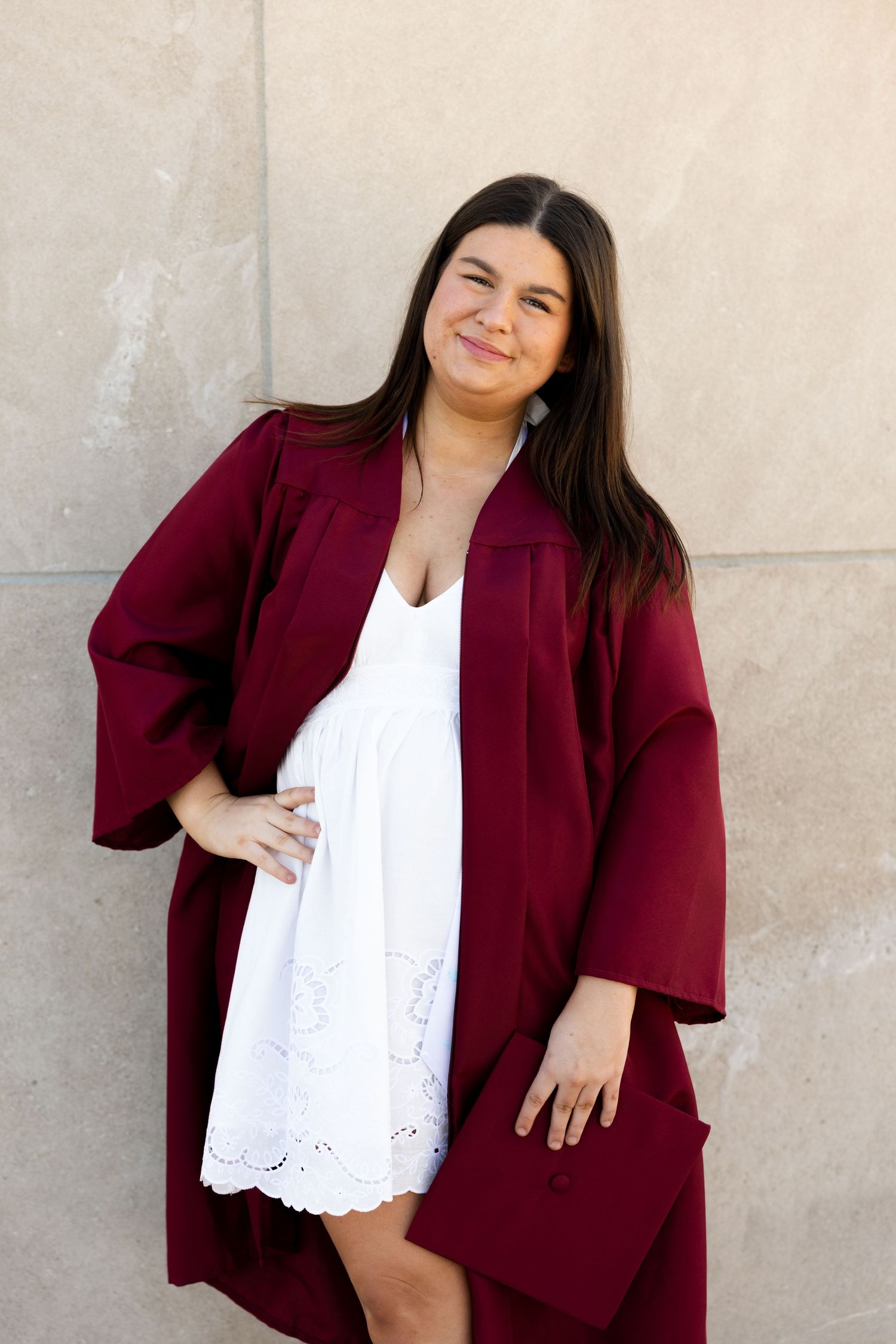Young woman smiling in maroon graduation cap and gown over white dress, posing outdoors against a stone wall.
