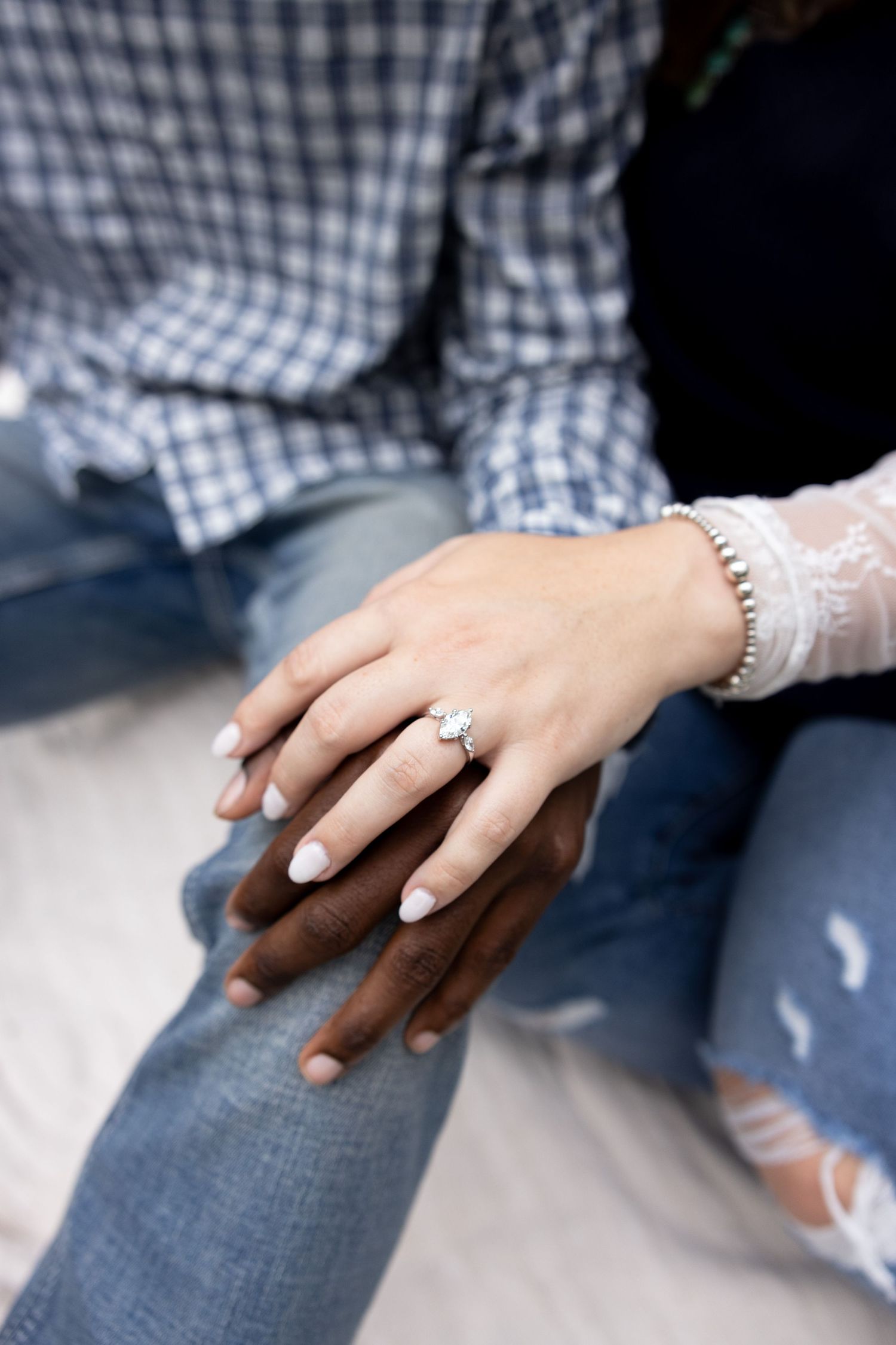 Two hands rest together showing an engagement ring, with one wearing a checkered shirt and the other in ripped jeans.