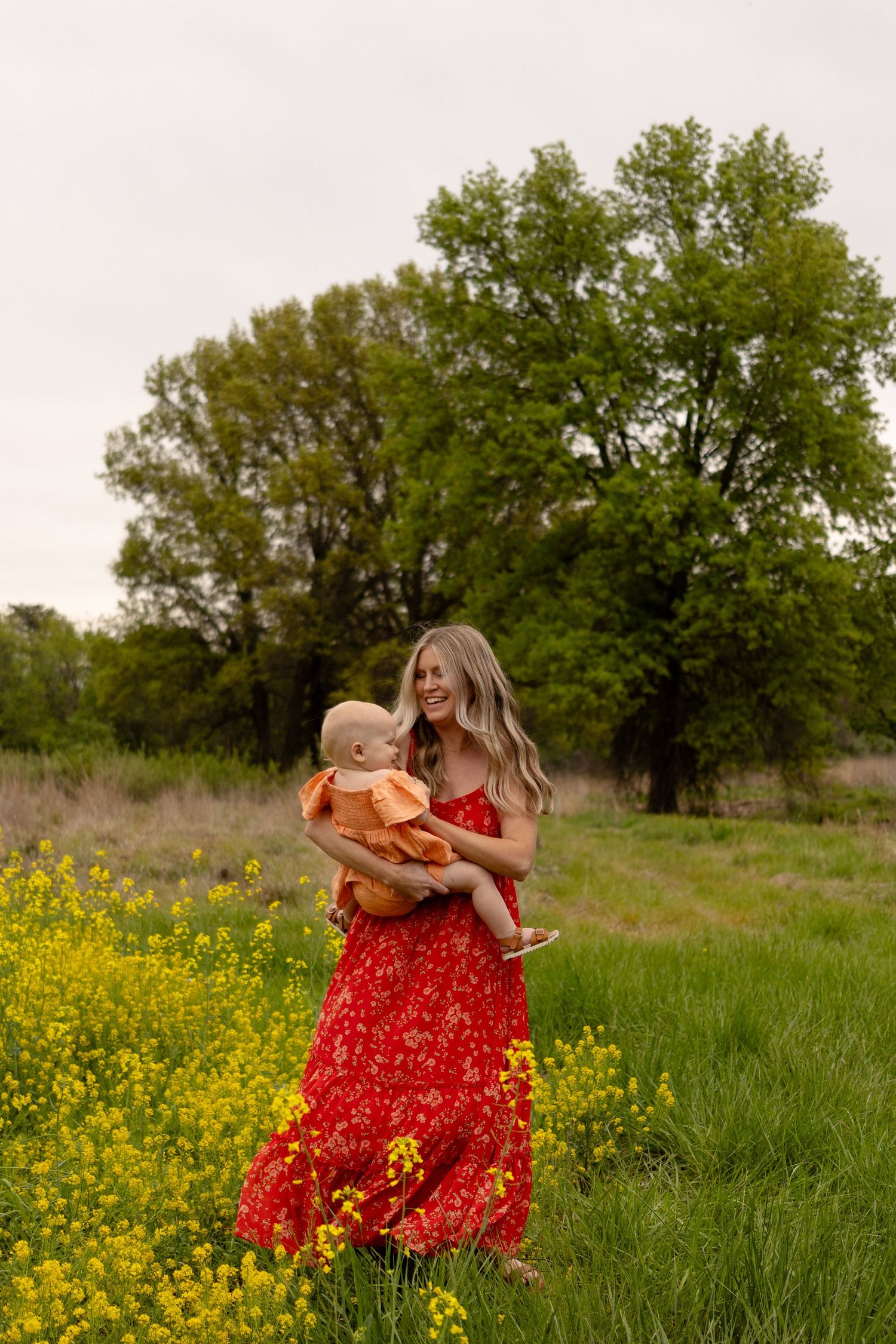 Woman in flowing red dress holds baby while standing in field of yellow wildflowers at sunset.