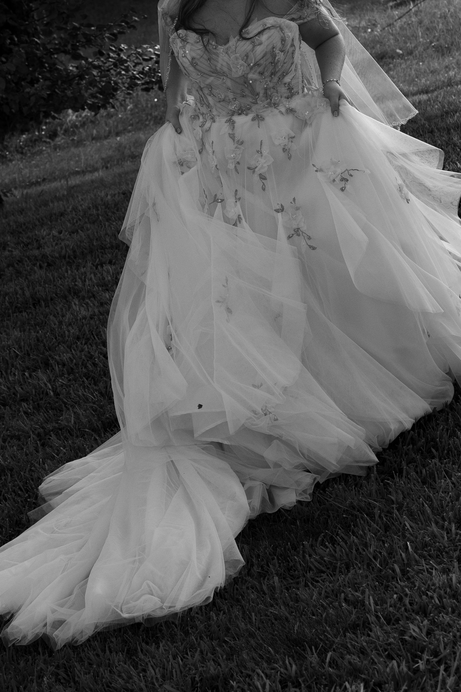 Black and white photo of a bride in a flowing tulle wedding gown with floral appliques and a long train outdoors.