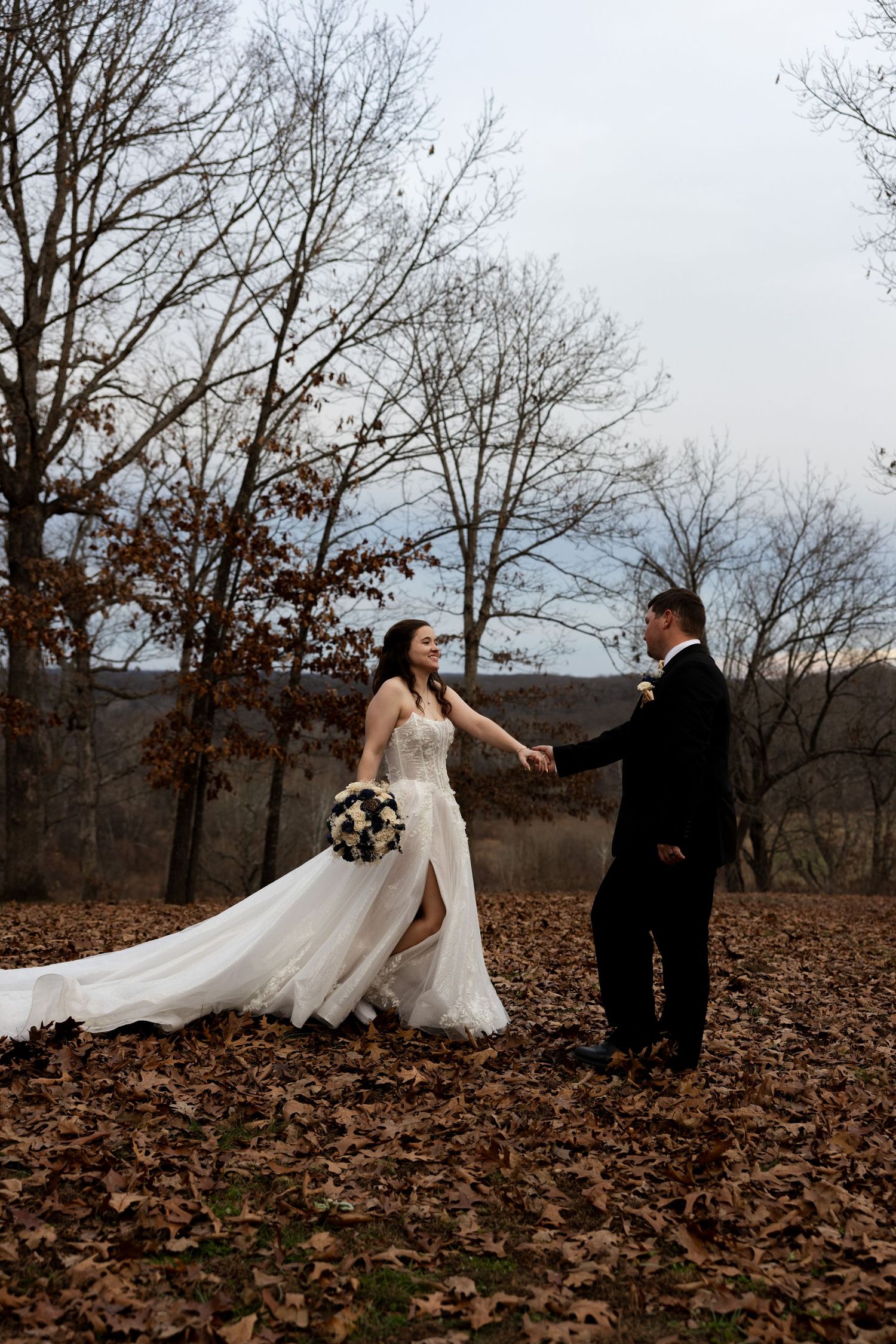 A romantic couple in wedding attire dances together on fallen autumn leaves in a wooded area during a fall photoshoot.