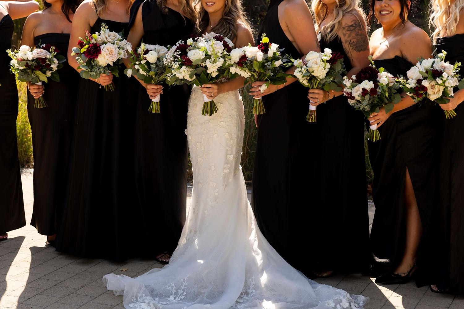 Wedding party in black dresses poses with bride holding white bouquets against dark background.