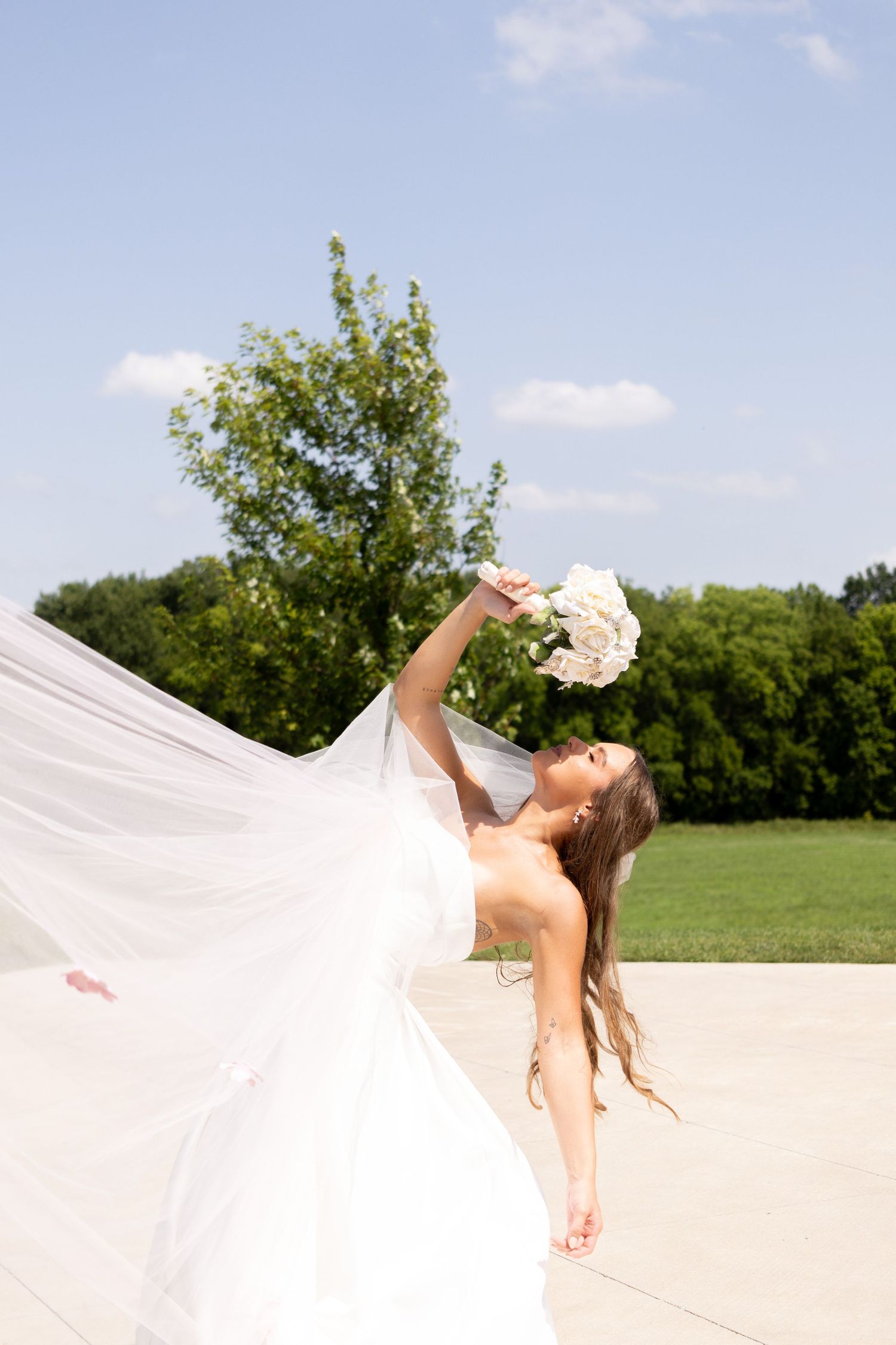 Bride in flowing white dress twirling gracefully in an open field against blue sky.