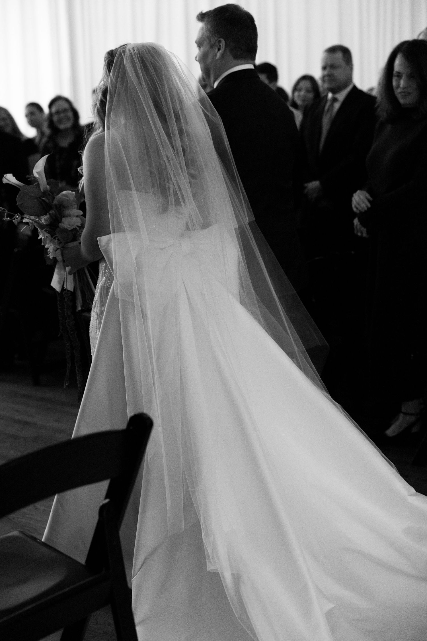 Black and white photo of bride in flowing gown and veil walking down the aisle with groom at wedding ceremony.