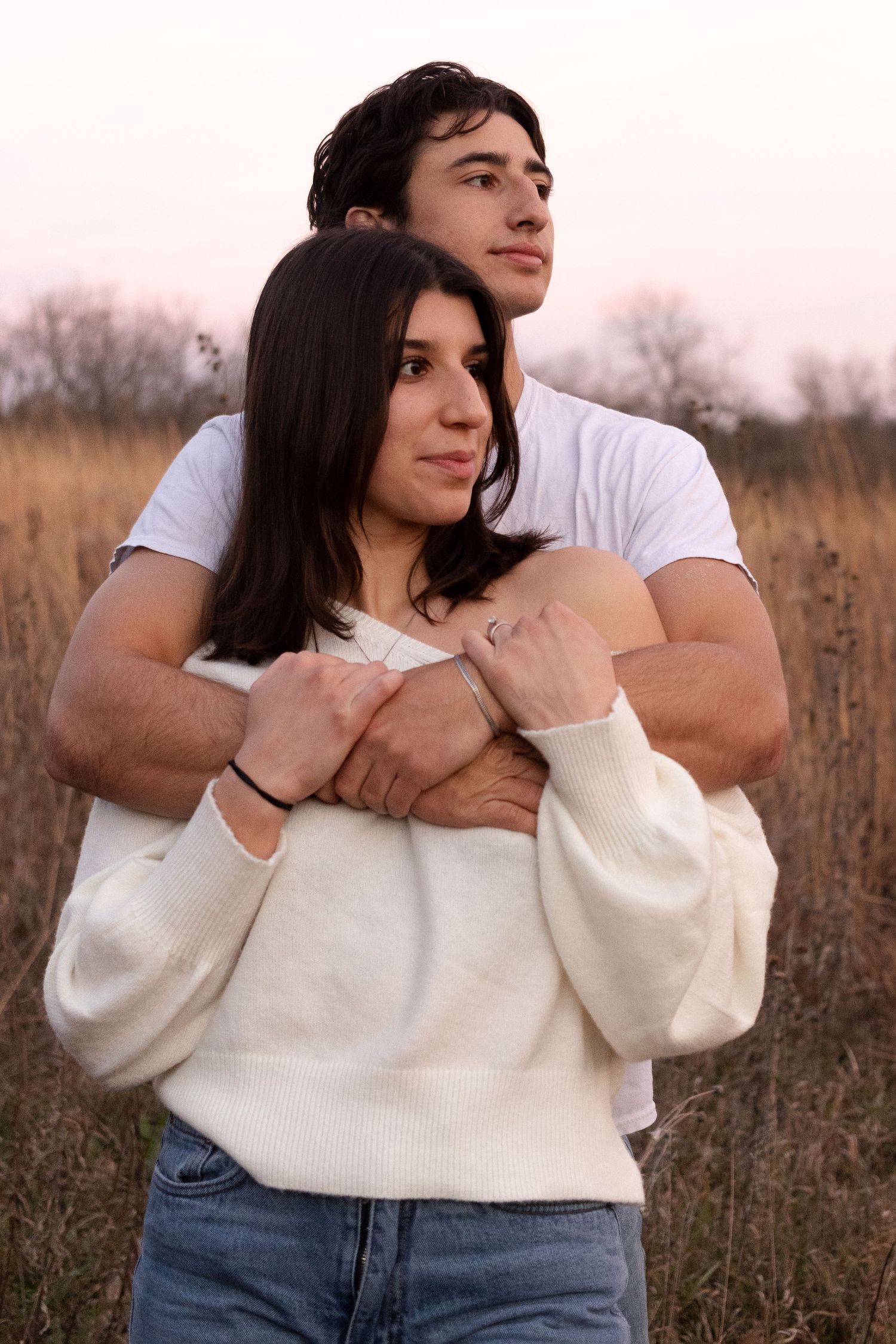 A couple embraces in a field during golden hour wearing white and casual clothing.