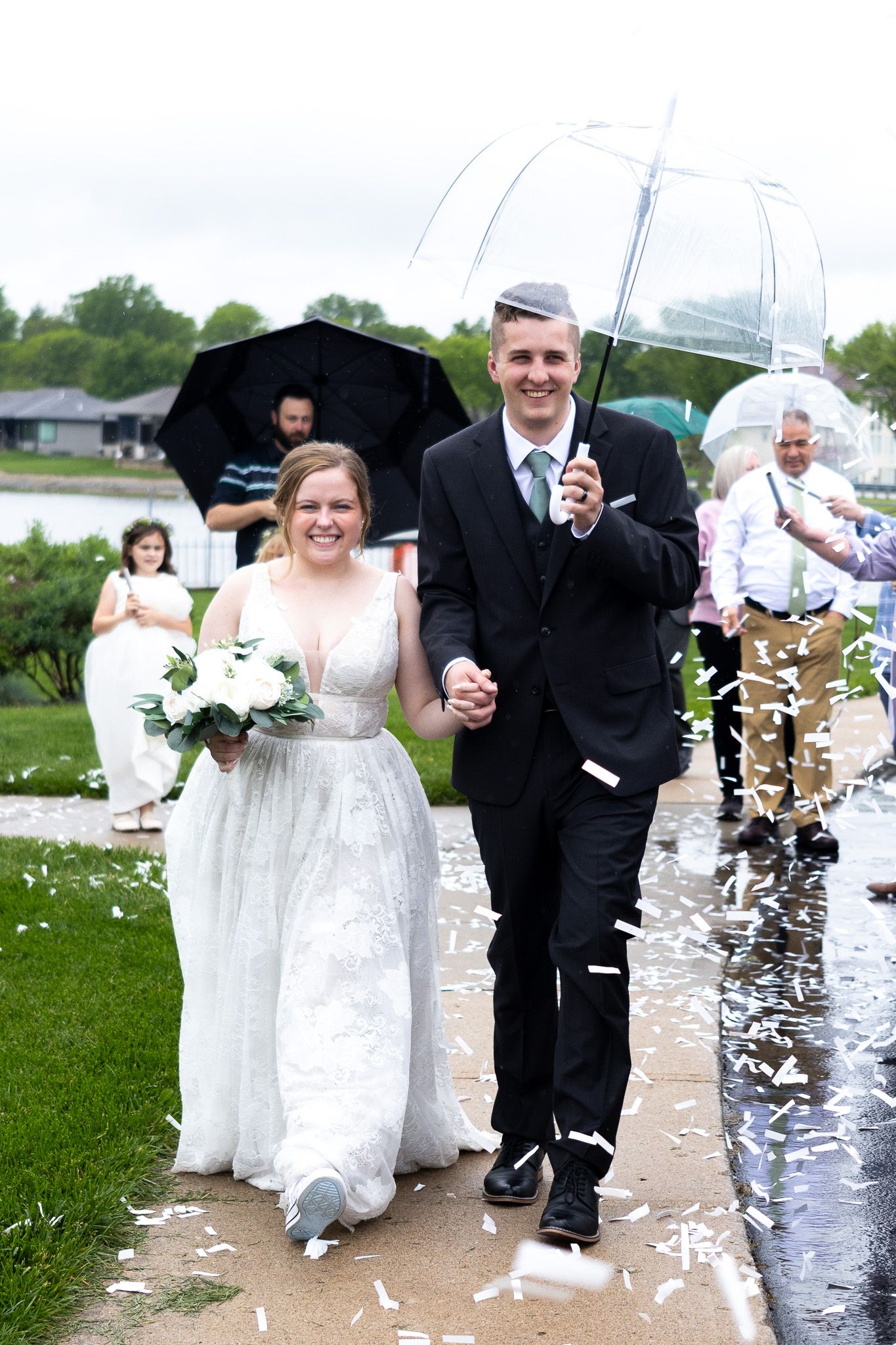 A wedding couple walks under umbrellas in the rain while confetti falls around them.