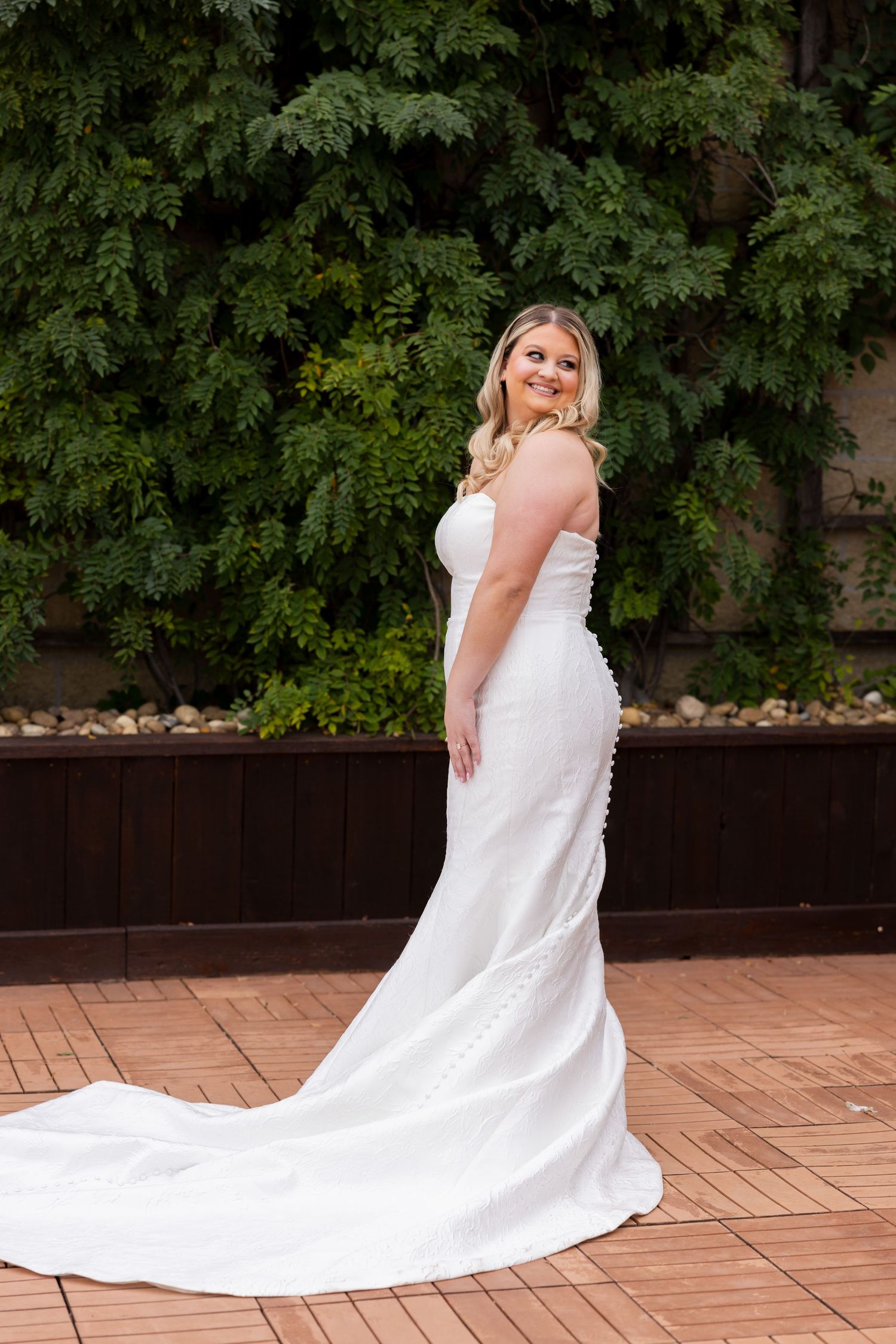 A person in an elegant white wedding gown with a long train poses on a brick patio against lush green foliage.