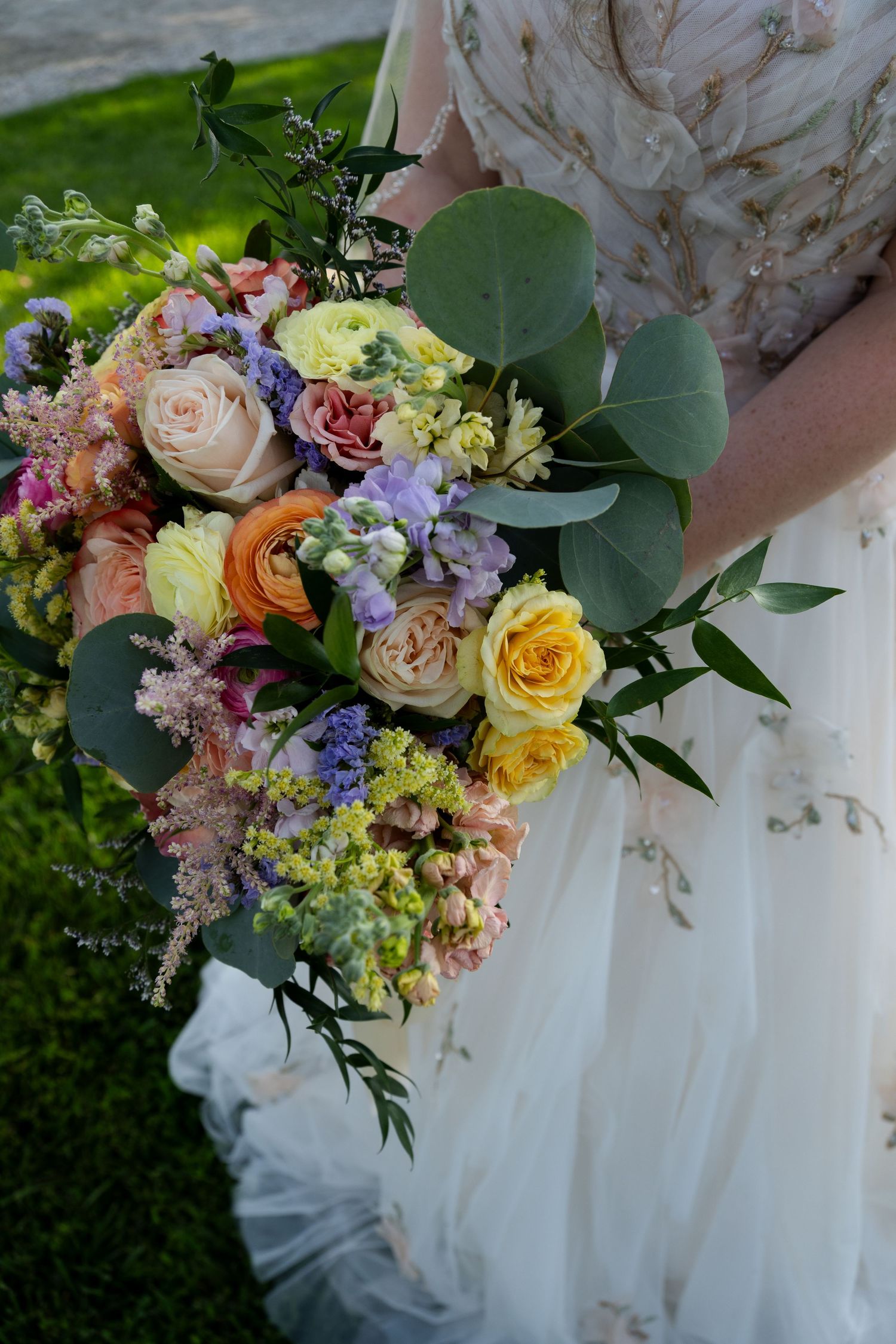 Bride holding a lush wedding bouquet with pastel roses, purple blooms, and eucalyptus greenery against a white gown.