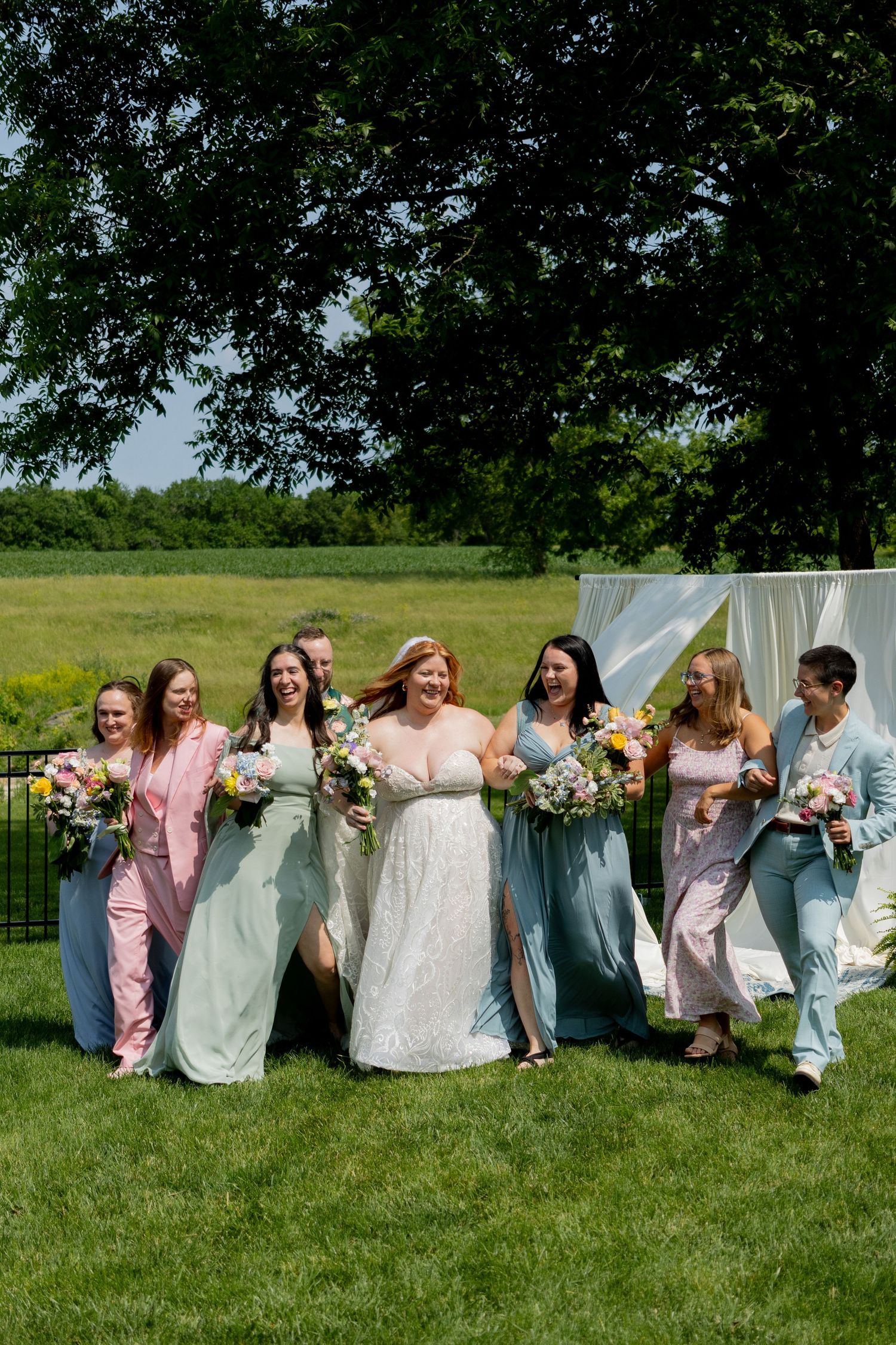 A group poses in pastel dresses against white hanging sheets on a sunny lawn.