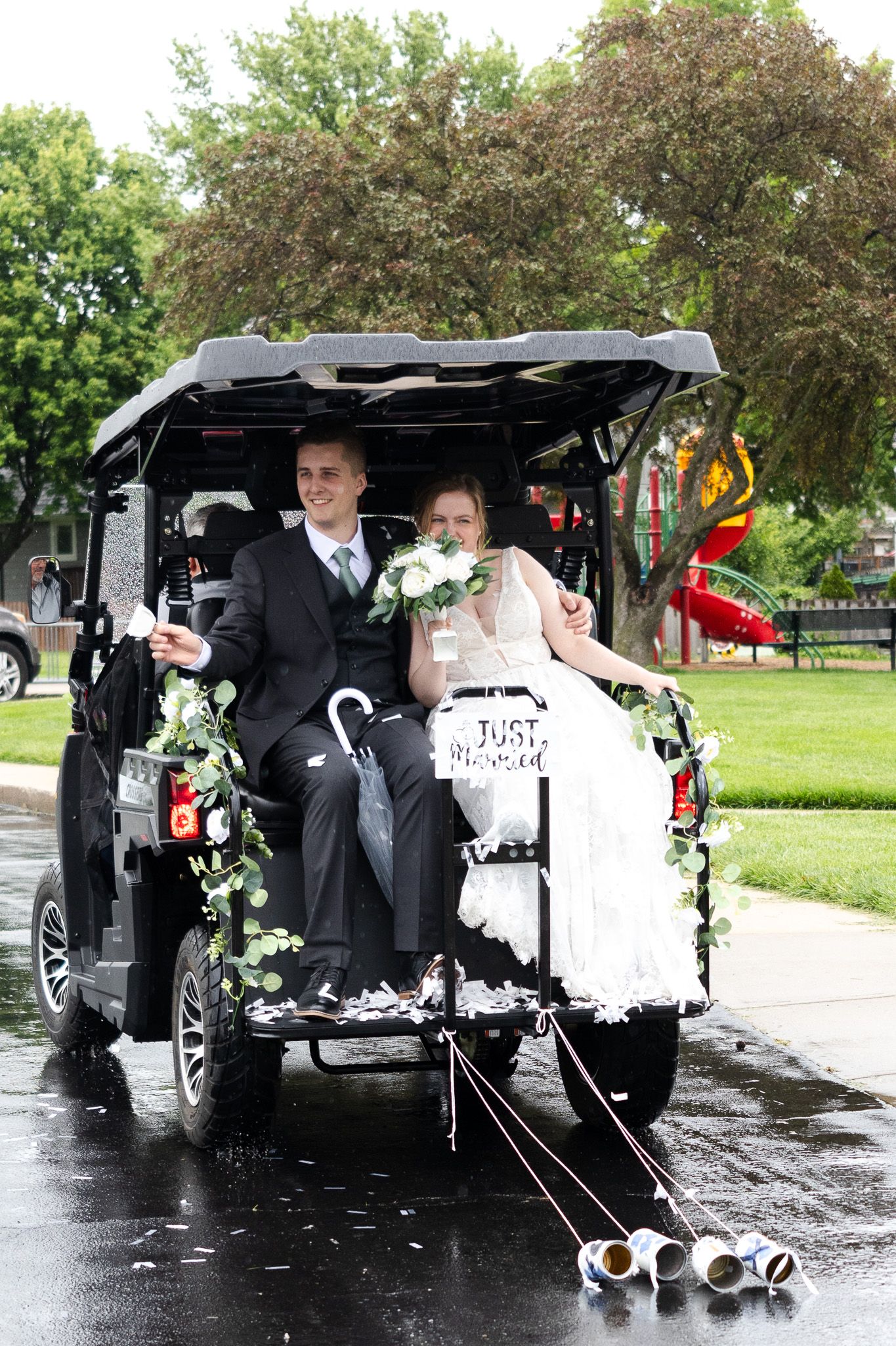 Newlyweds sit in a decorated golf cart with Just Married sign and tin cans trailing behind.