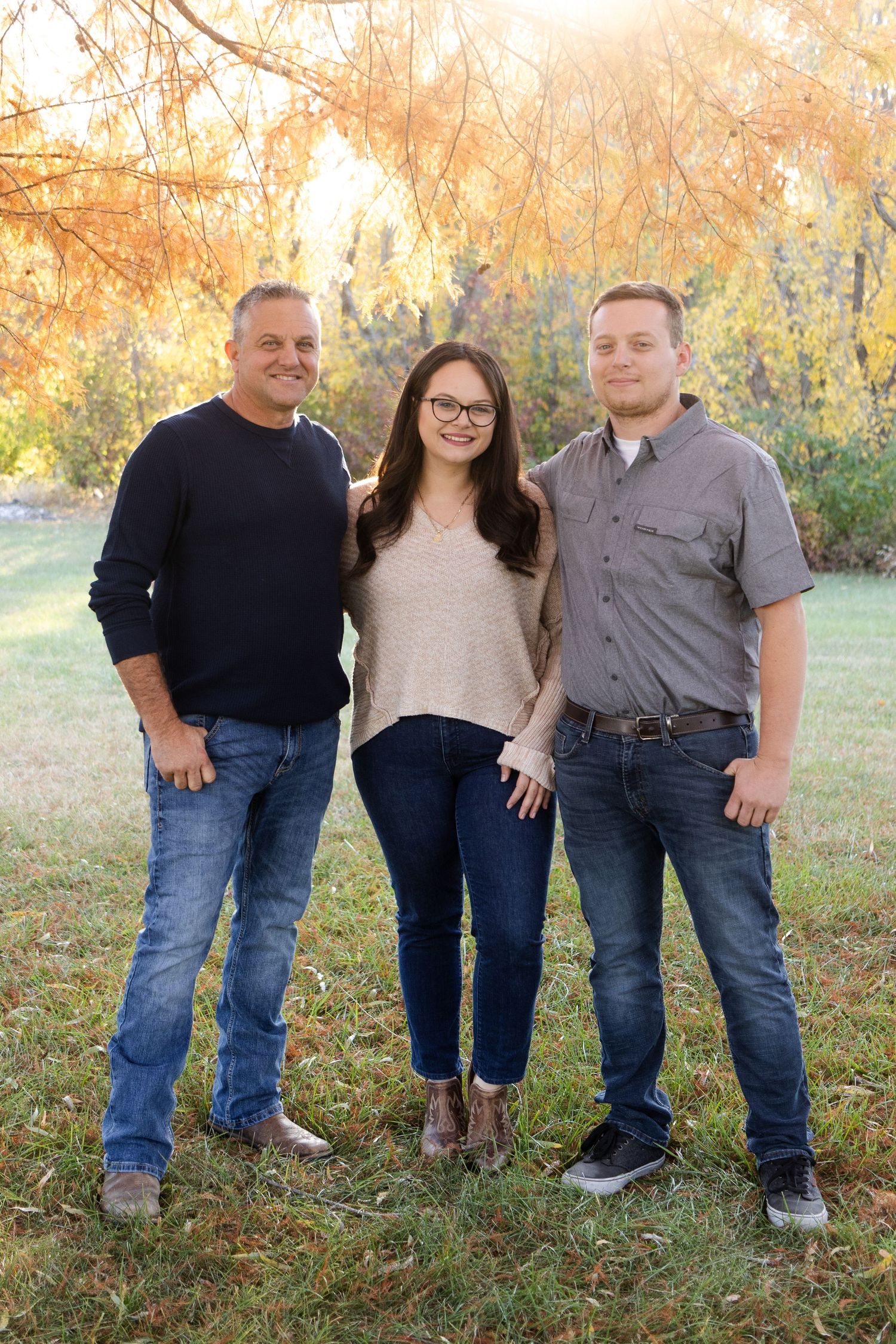 Three people stand together outdoors with autumn foliage in the background during a casual family photoshoot.