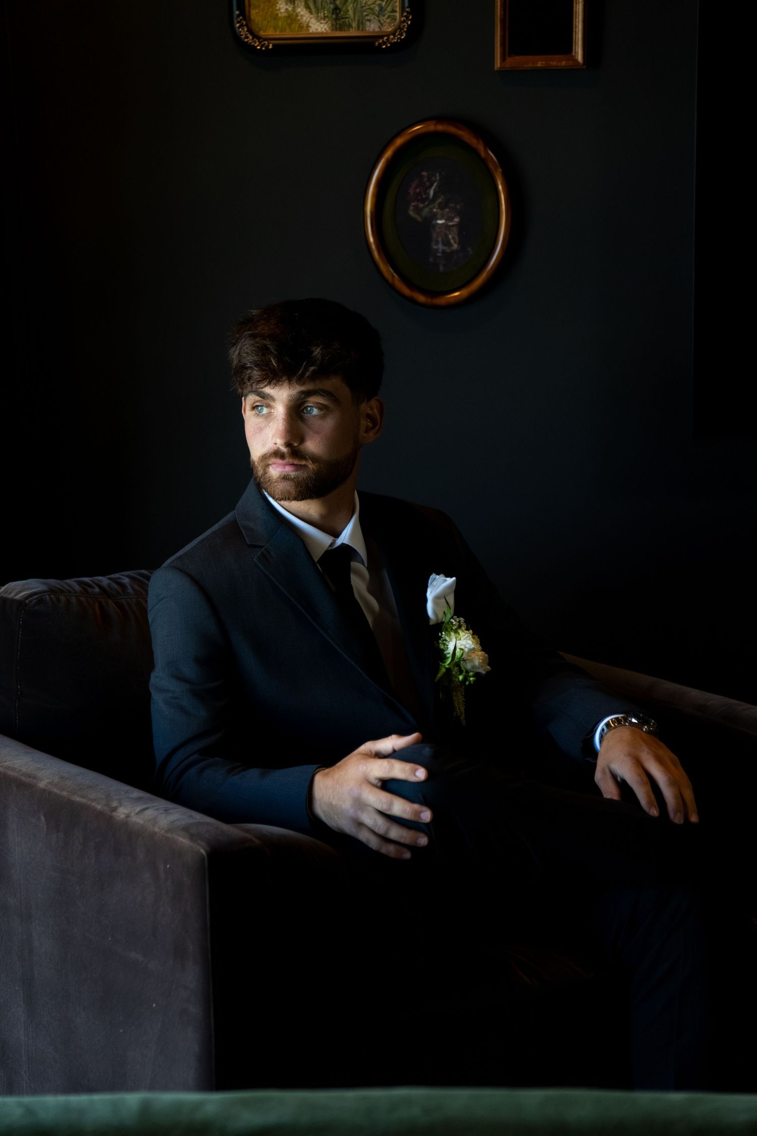 A dapper groom in a dark suit with boutonniere sits pensively on a velvet sofa against a moody dark wall with framed art.