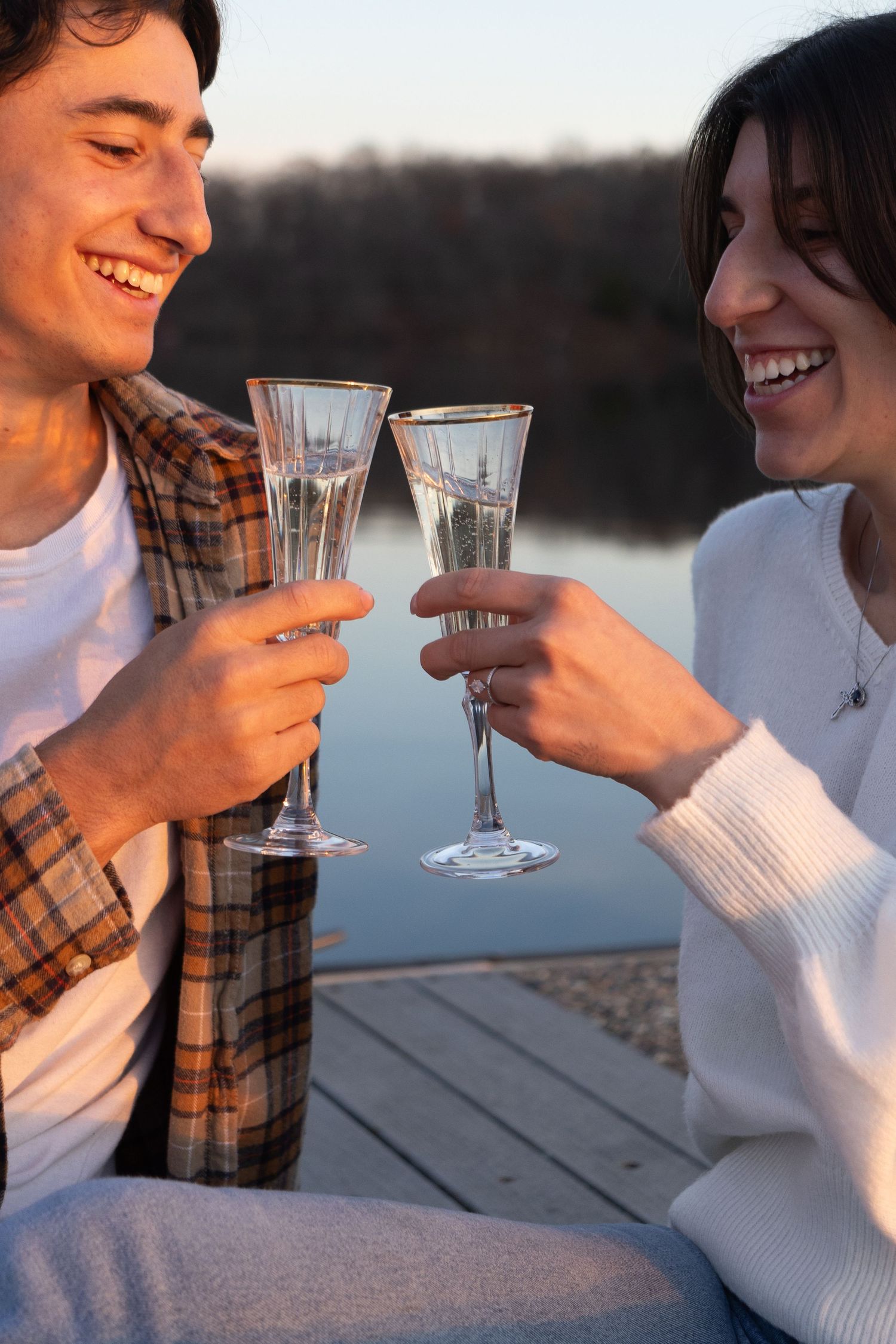 A couple shares a romantic toast with champagne glasses while sitting on a dock at sunset by the water.