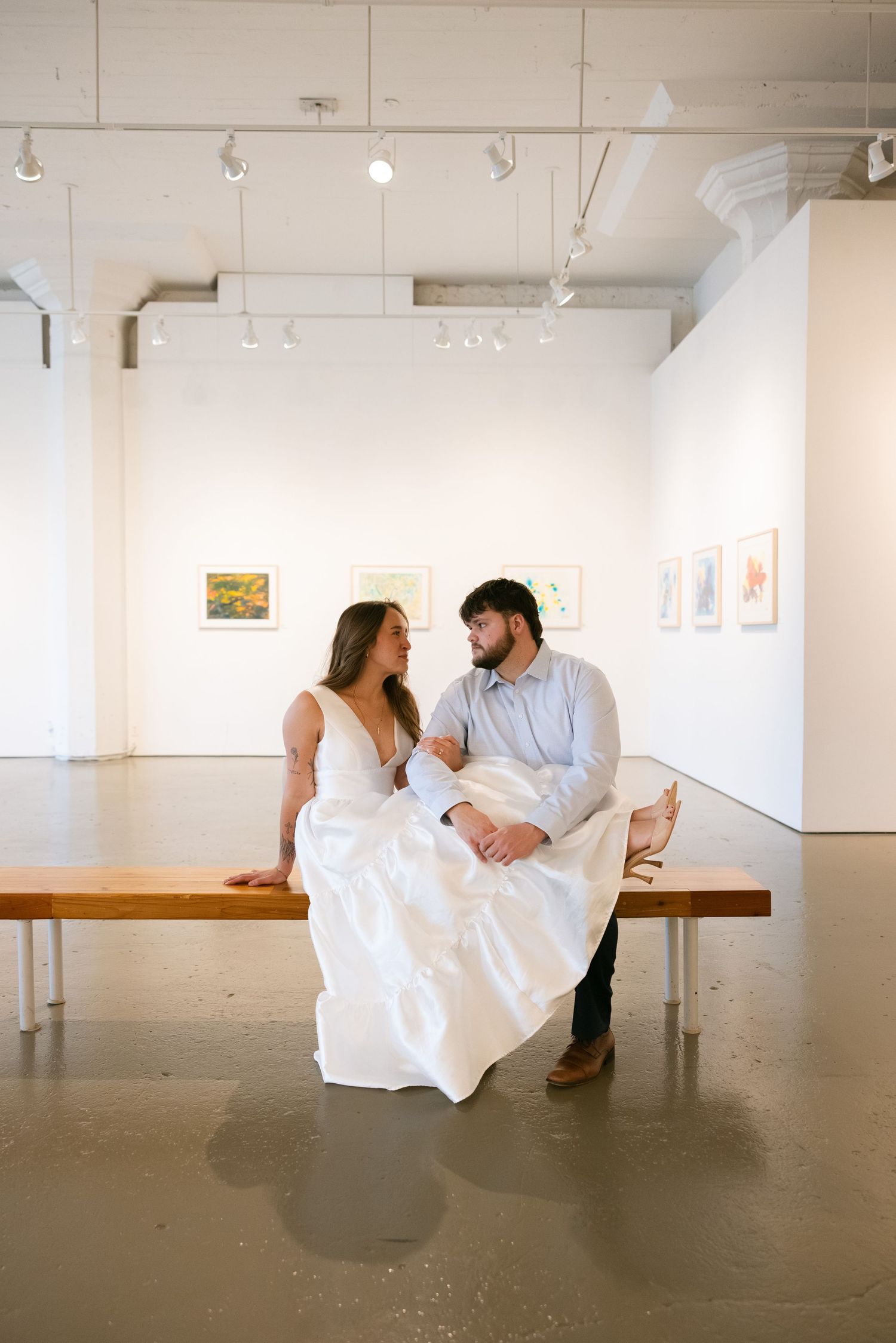 Bride and groom sitting on a wooden bench in an art gallery, gazing at each other lovingly in white wedding attire.