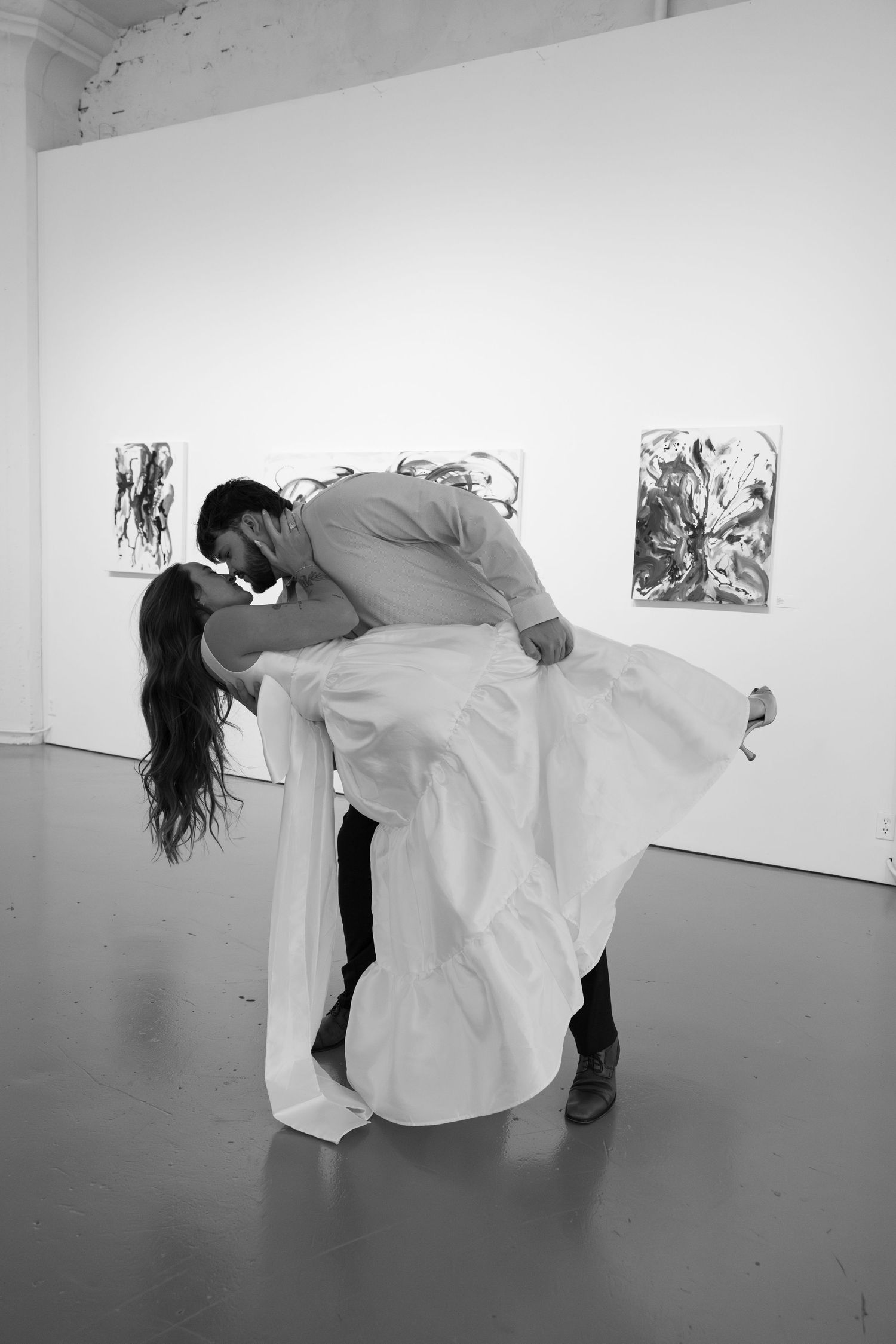 Black and white photo of a couple sharing a romantic dip kiss in an art gallery, bride in flowing white gown.
