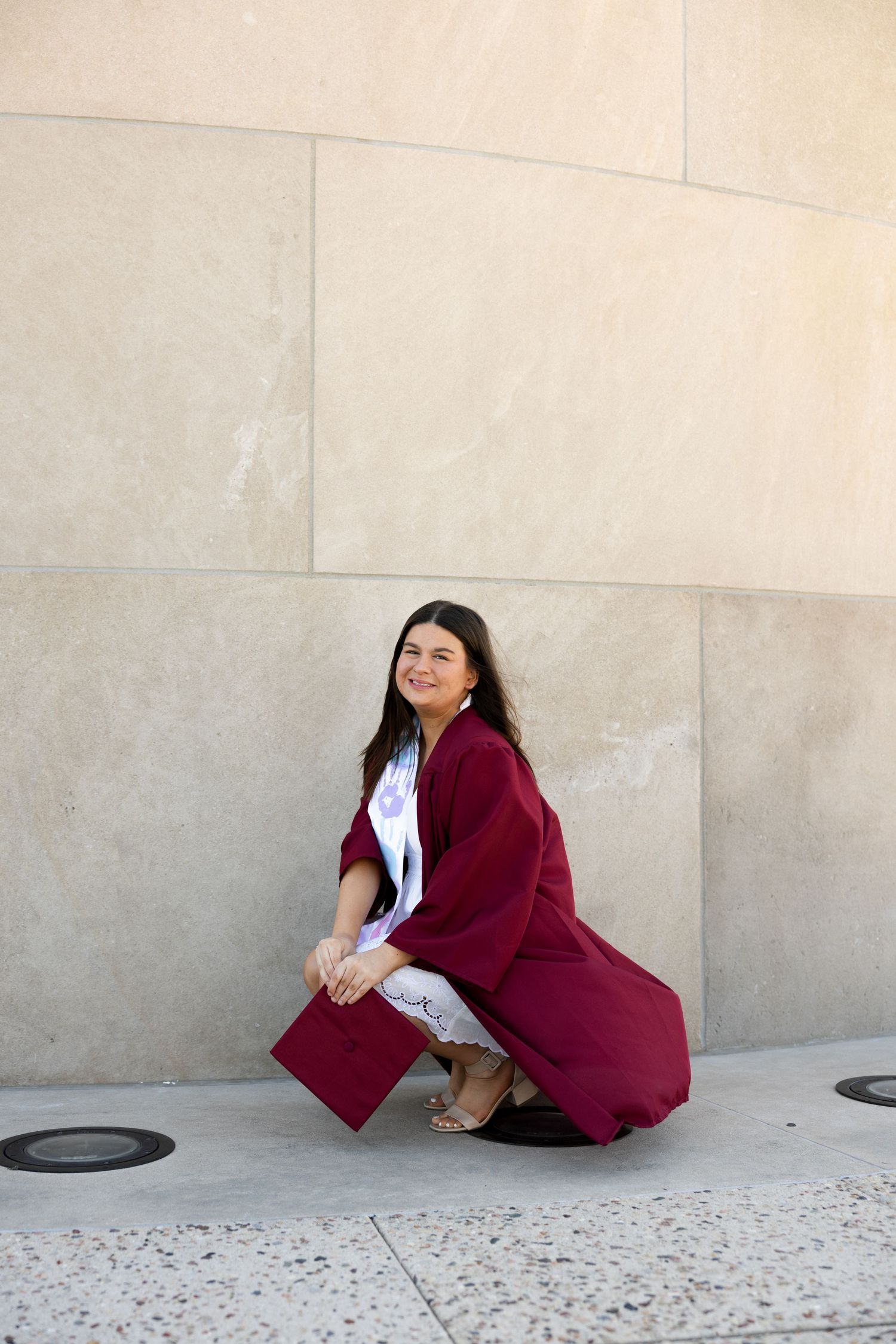 Young woman in maroon graduation gown and cap crouching outdoors, smiling over shoulder near stone wall.