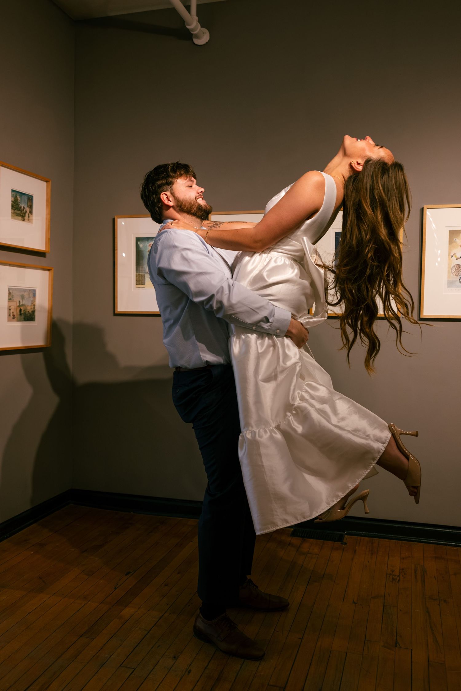 A couple dances playfully in an art gallery space with wooden floors and framed artwork on display.