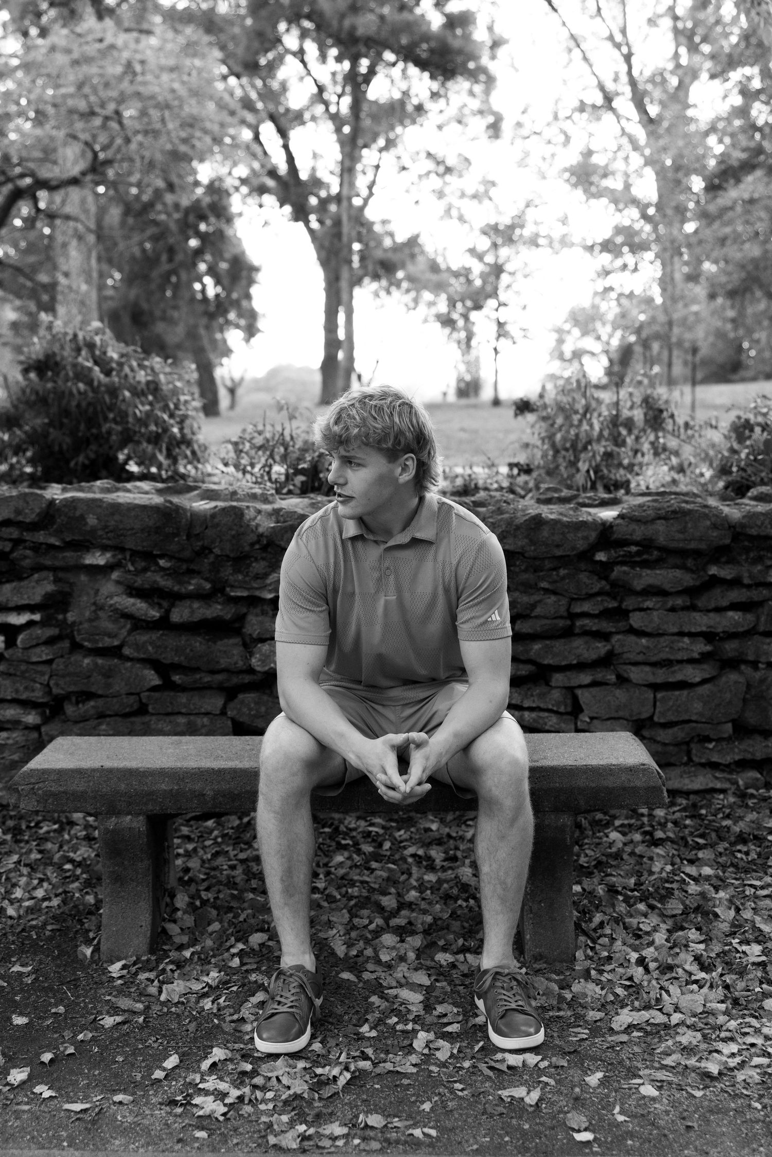A person sits alone on a stone bench in a park setting with trees and a stone wall in black and white photography.