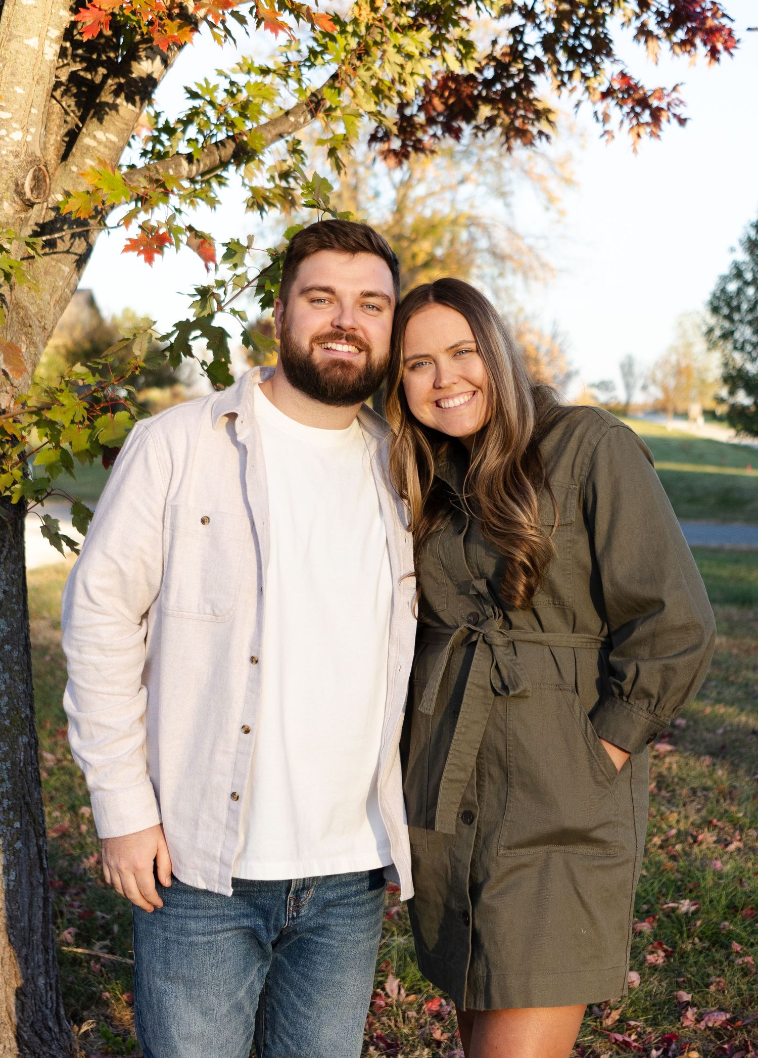 Couple poses together under colorful autumn tree branches in casual attire.