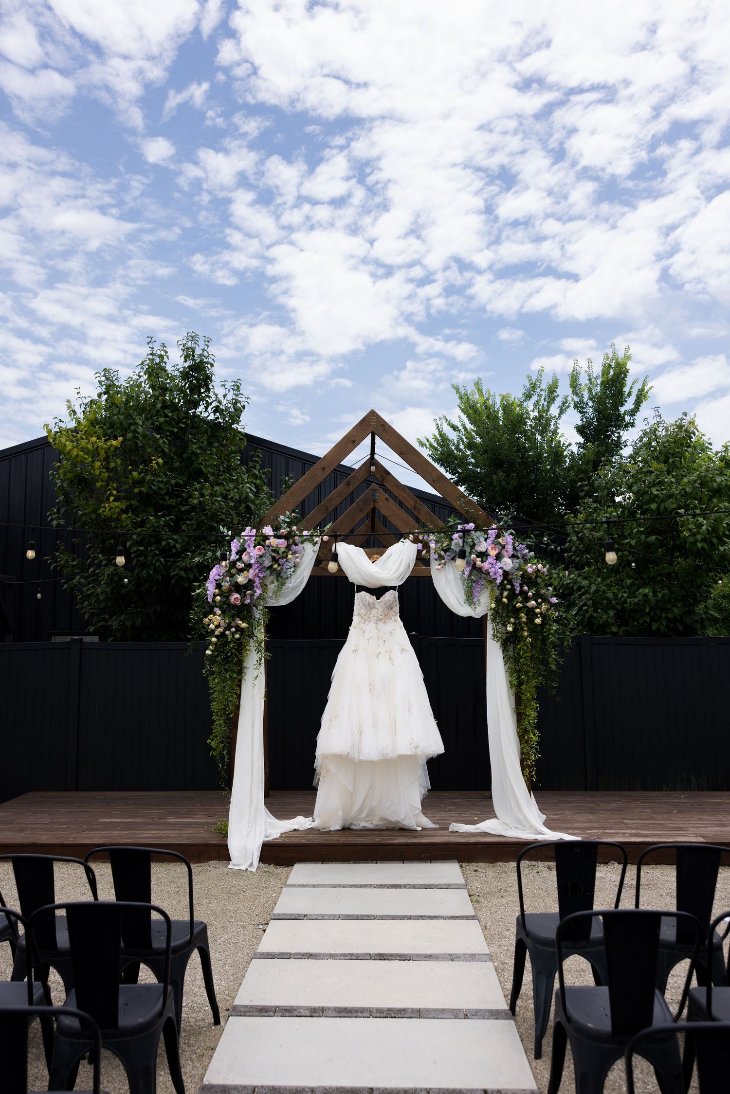 Elegant outdoor wedding ceremony setup with wooden arch, floral decorations, white draping, black chairs, and a bridal gown on display.