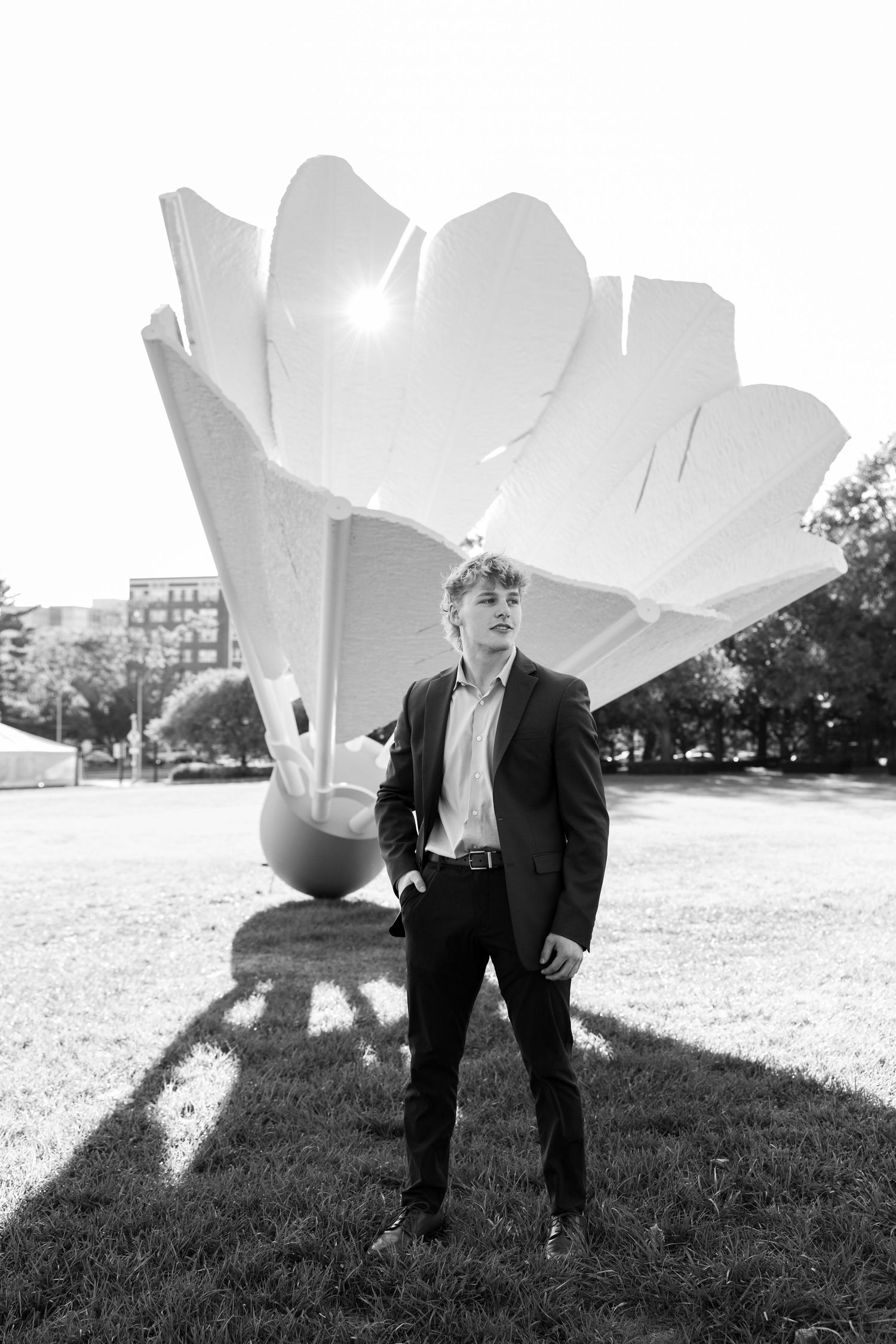 A figure in a dark suit stands in a grassy field with large white sculptural flower petals in the background.