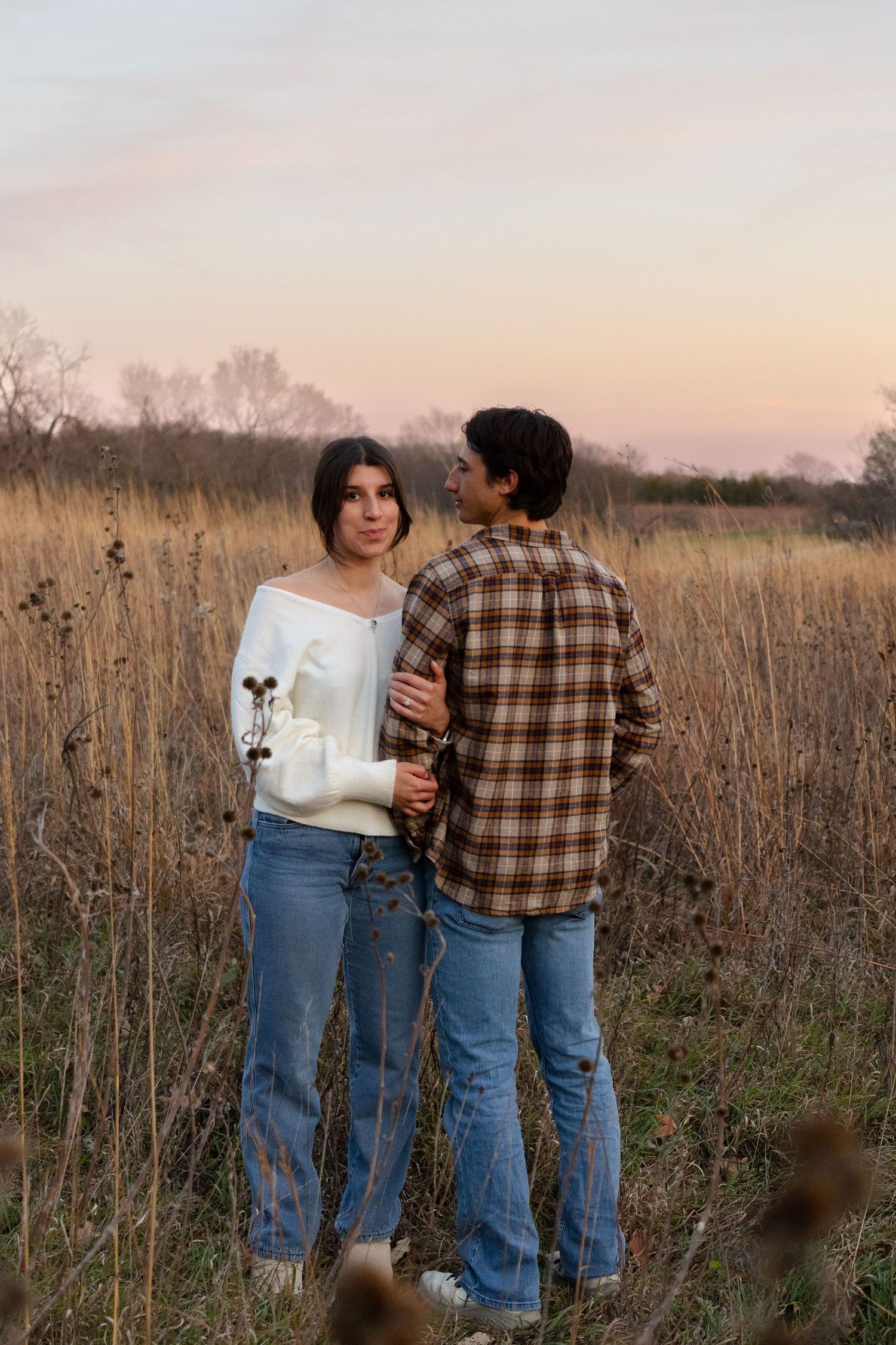 A couple in casual wear standing together in a field of tall grass at sunset.