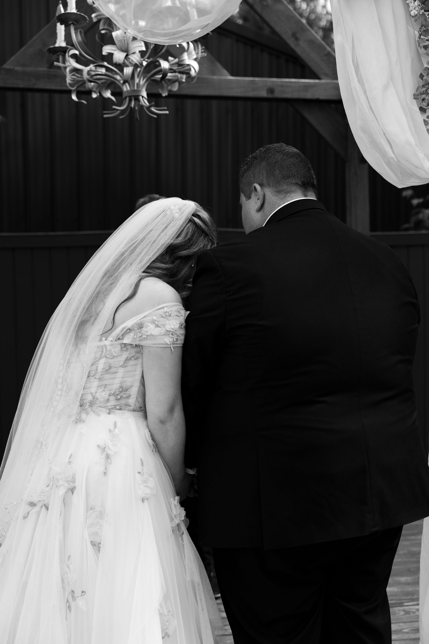 Black and white photo of bride and groom seen from behind at outdoor wedding ceremony with chandelier.