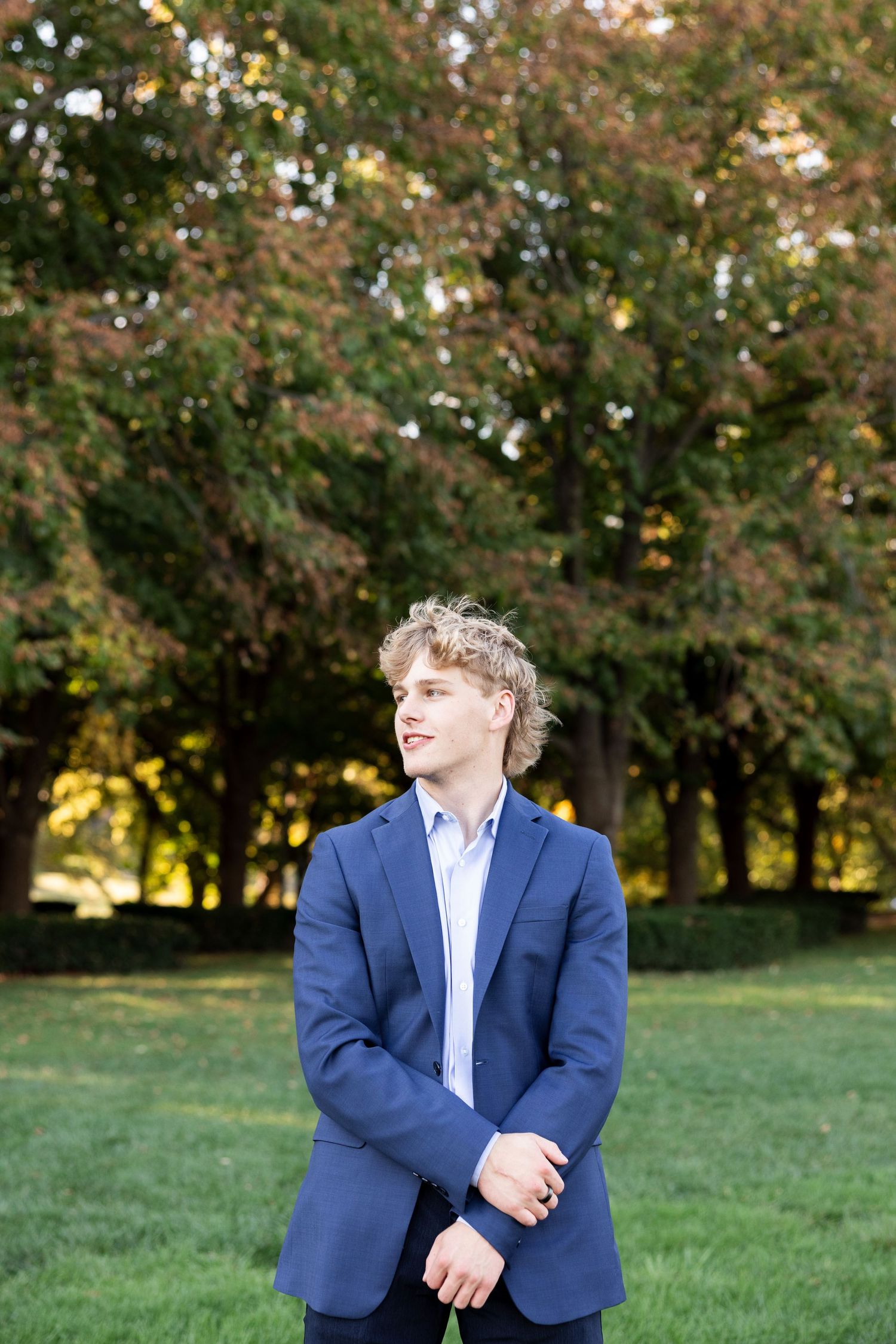 A figure in a navy blue suit stands in a grassy outdoor setting with autumn trees in the background.