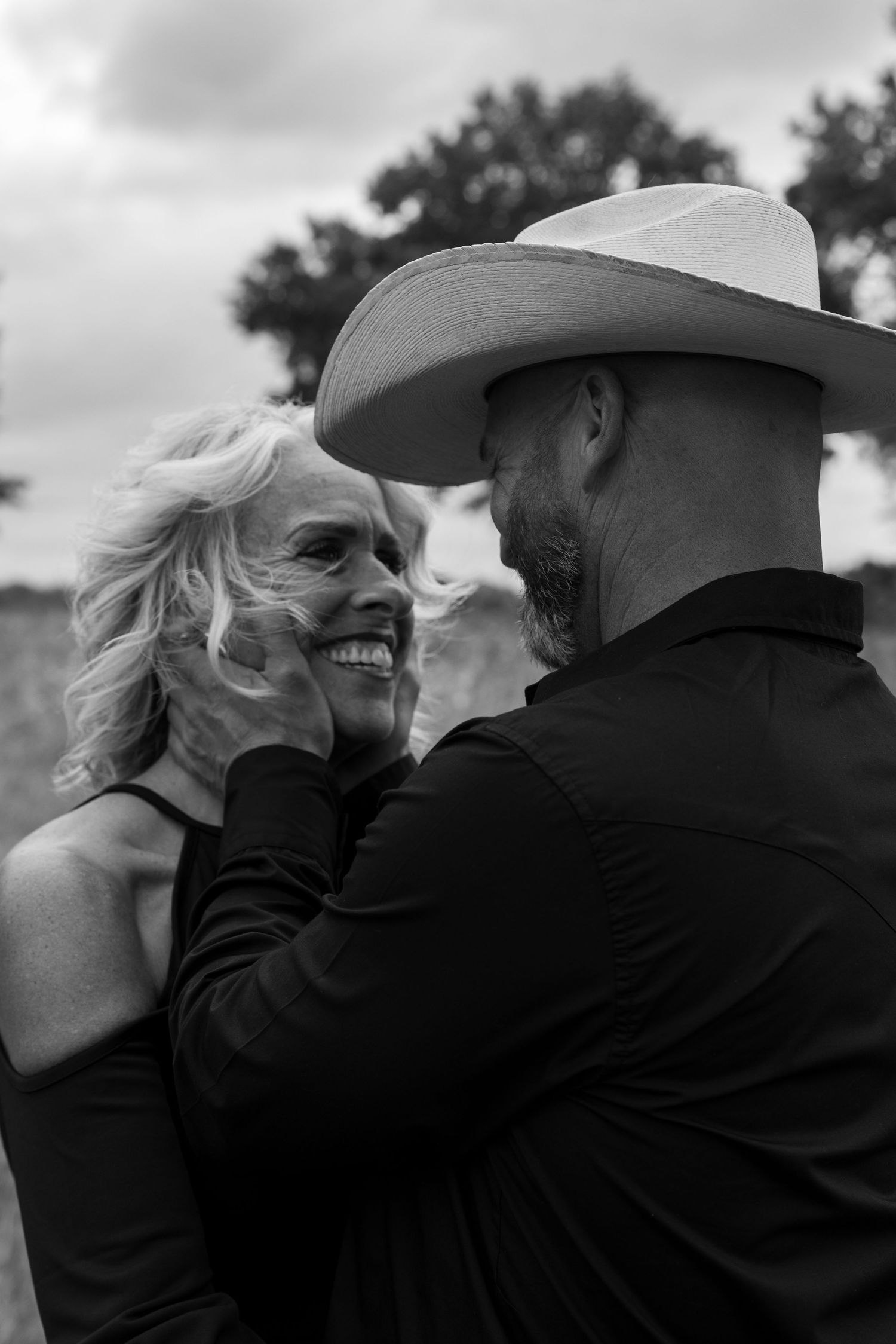 Black and white photo of a smiling woman and a man in a cowboy hat sharing a tender moment outdoors.