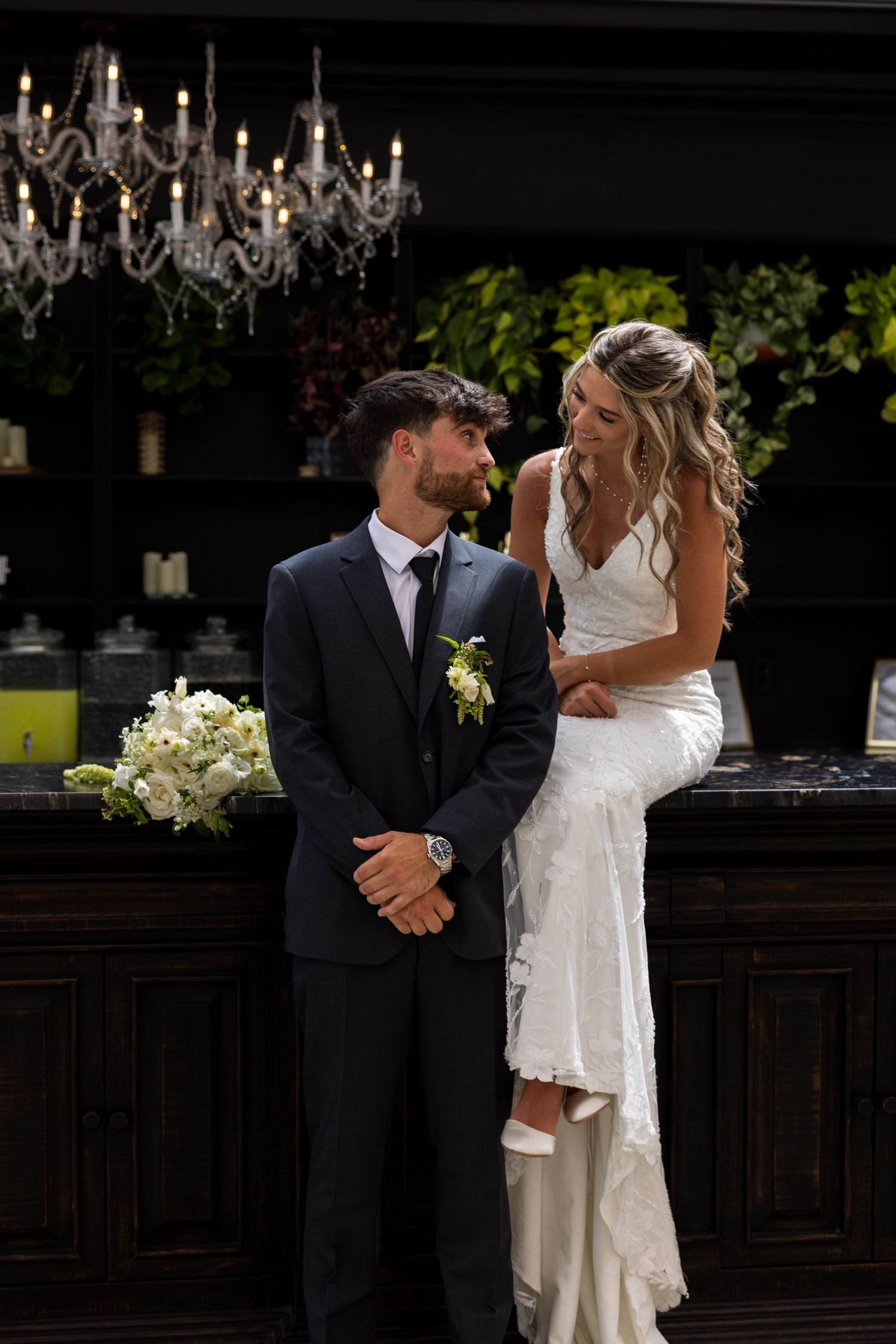 Bride in white lace gown sits on bar counter gazing at groom in dark suit with boutonniere at elegant wedding venue.