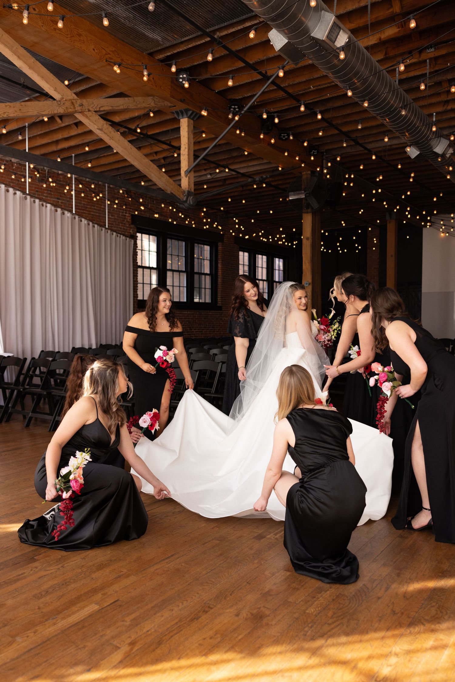 Bridesmaids in black gowns help arrange bride's white wedding dress and veil in elegant string-lit venue.