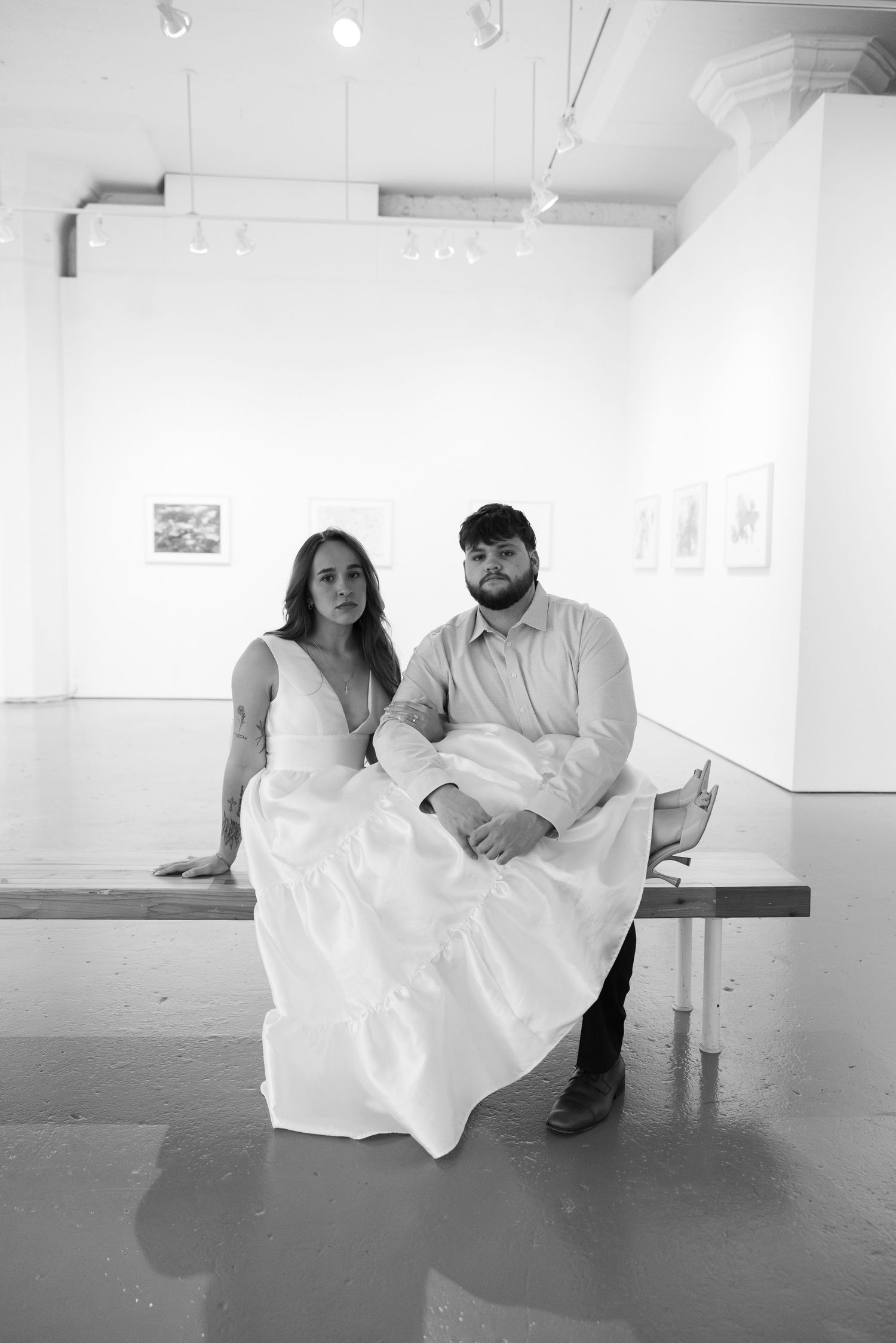 A couple sits together on a bench in a minimalist white gallery space, captured in black and white photography.