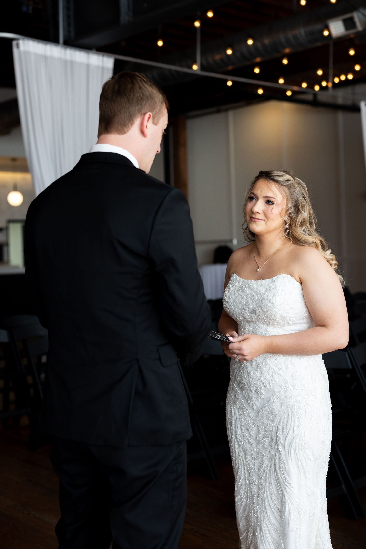 Bride in strapless beaded white gown smiles at groom in black tuxedo during wedding ceremony indoors.