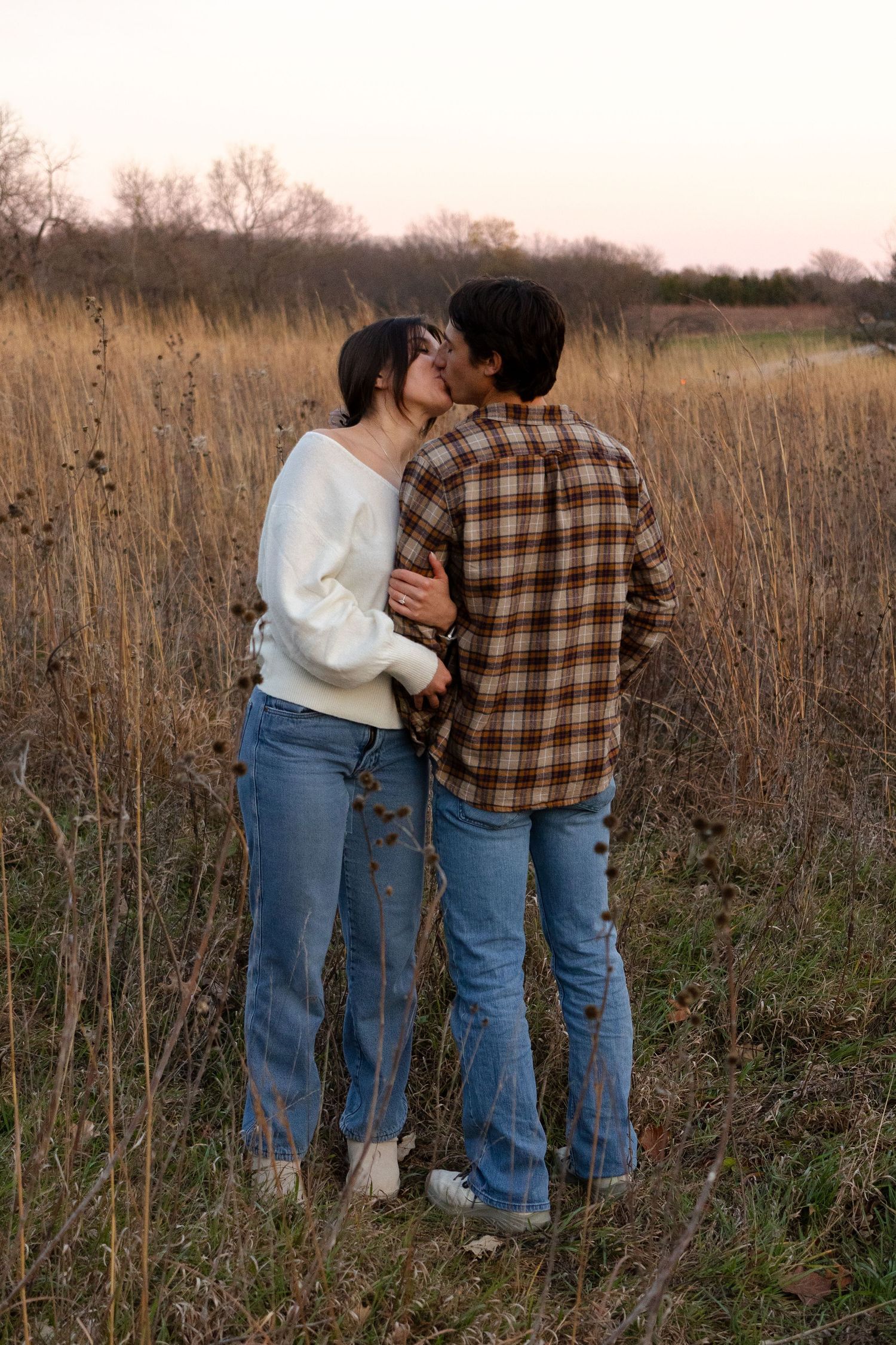 A couple in jeans and plaid shirt embrace in a rustic autumn field at sunset.