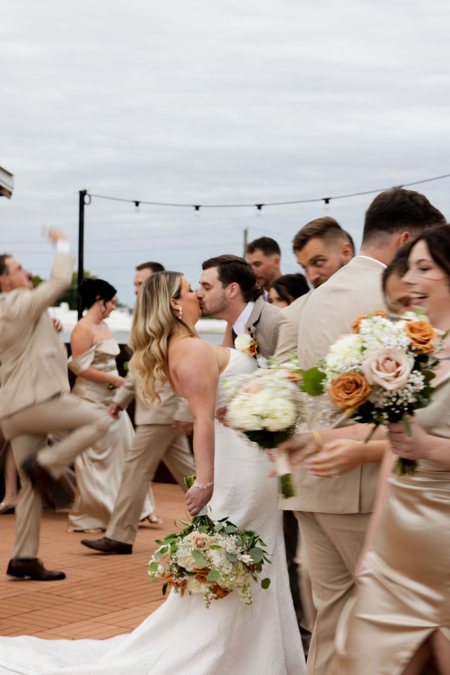 Wedding party in beige and neutral tones poses on a deck with floral bouquets against an overcast sky.