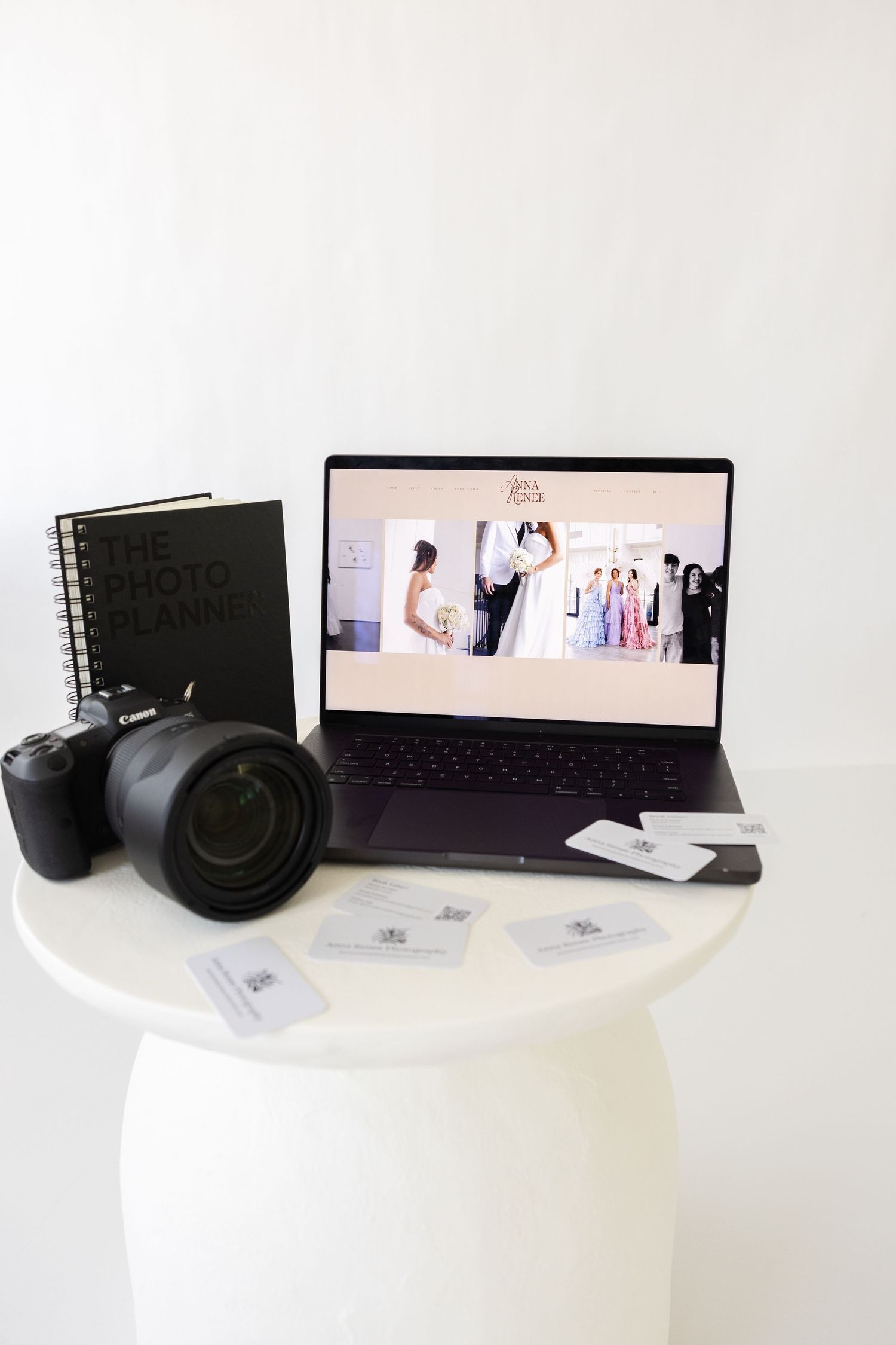 A Canon camera, laptop displaying a wedding photography website, business cards, and a photo album arranged on a white round table.