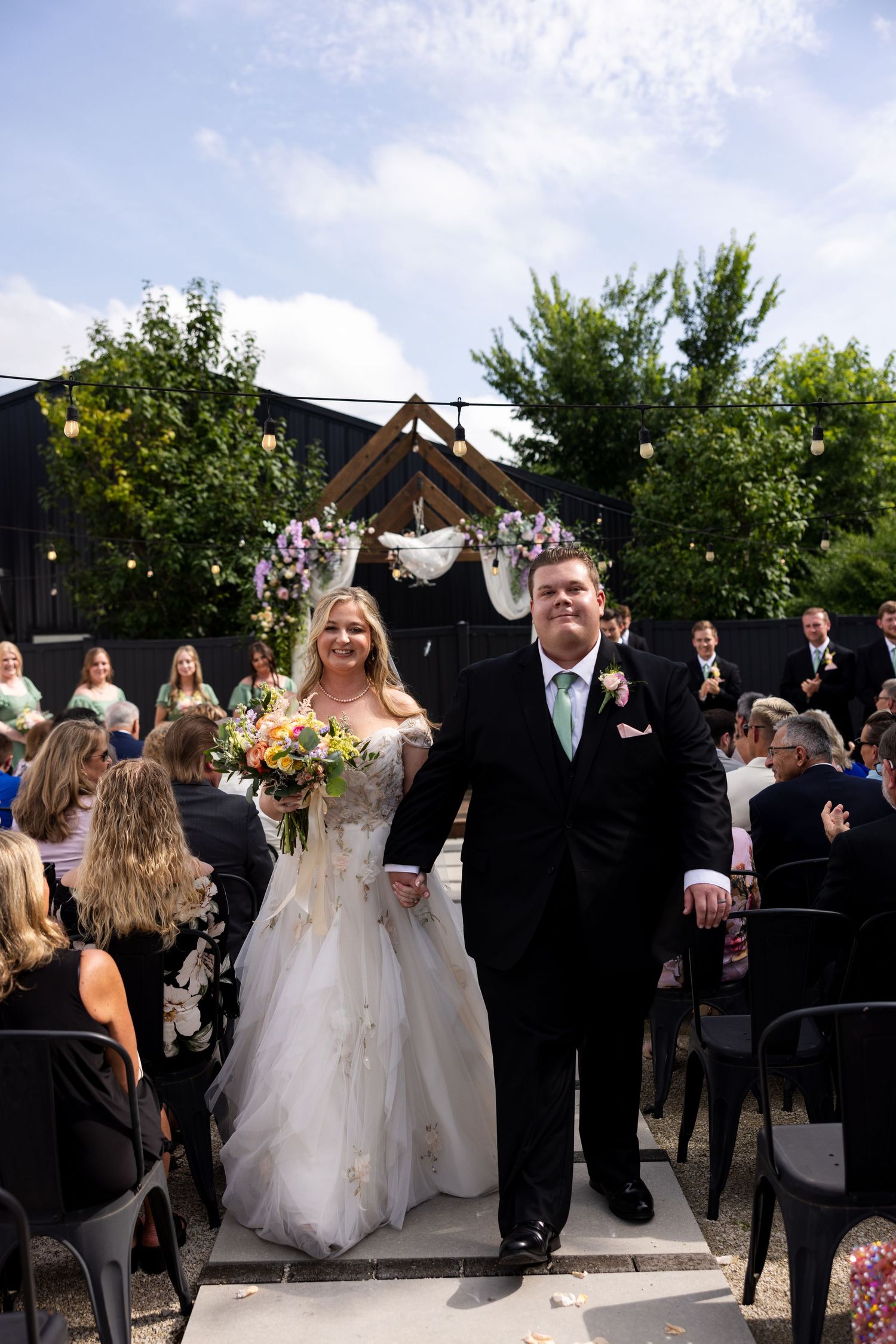 Bride and groom walk down outdoor wedding aisle, bride holding colorful bouquet, guests seated, floral arch backdrop.