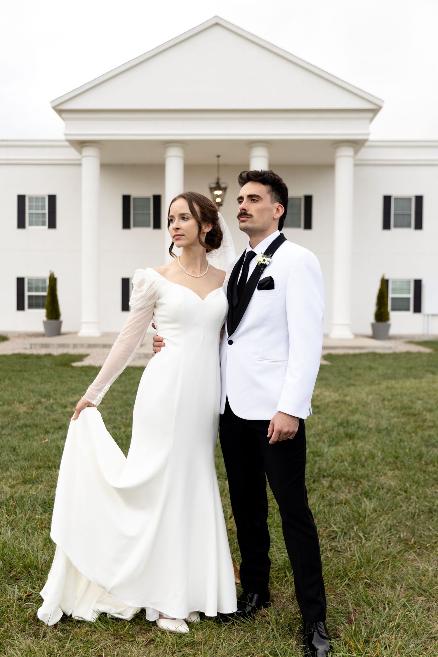 Elegant bride in white gown and groom in white tuxedo jacket pose before a grand white colonial mansion.