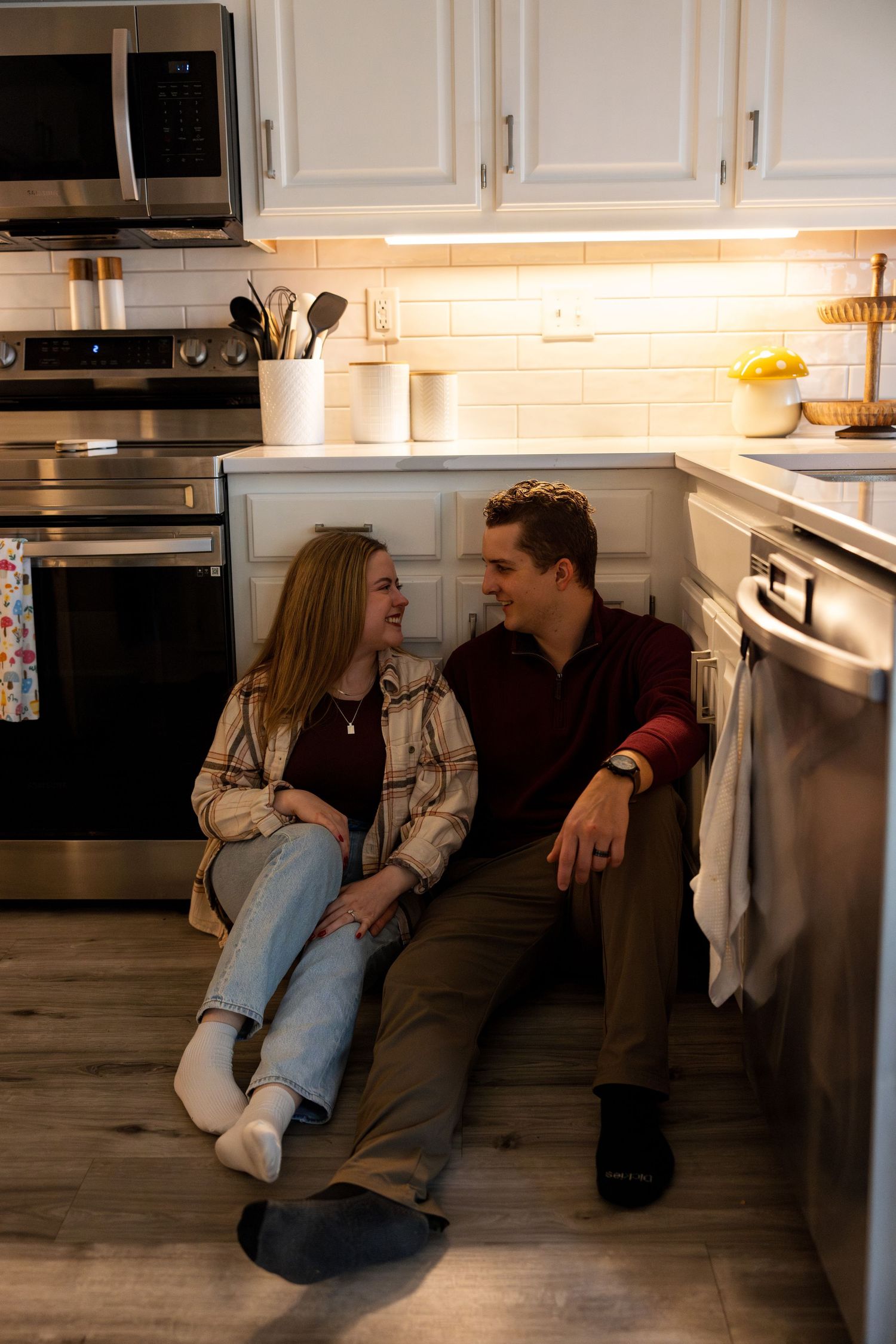 Two people sit and chat intimately on a kitchen floor with white cabinets.